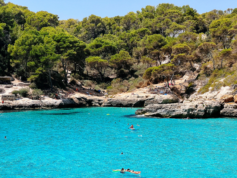 A scenic coastal cove featuring turquoise blue water with several people swimming and kayaking. The rocky shoreline is bordered by lush green trees and vegetation. Several individuals can be seen relaxing on the rocks near the water&rsquo;s edge.