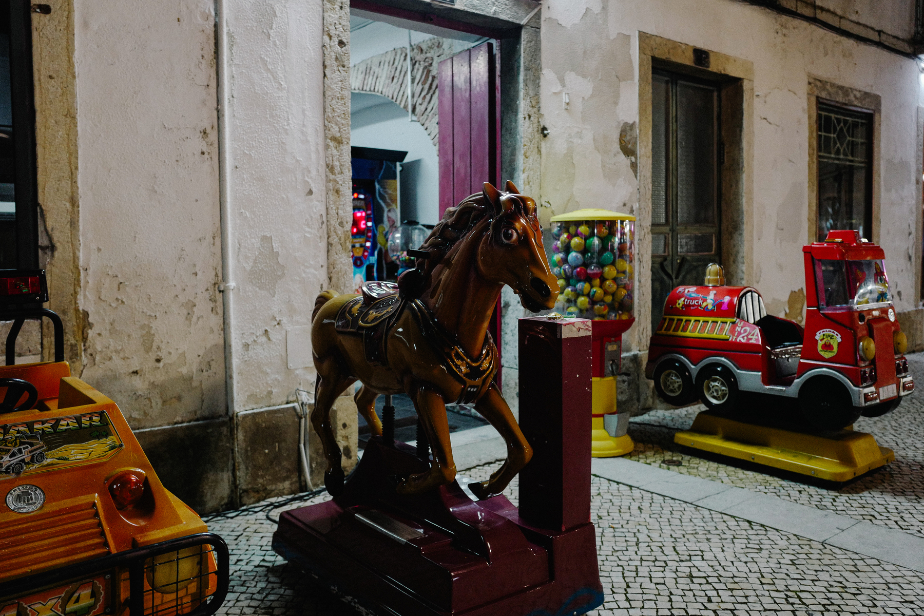 A coin-operated ride-on horse, a mini off-road vehicle, and fire truck for children beside a gumball machine, set on a cobblestone street against an old building with peeling paint.