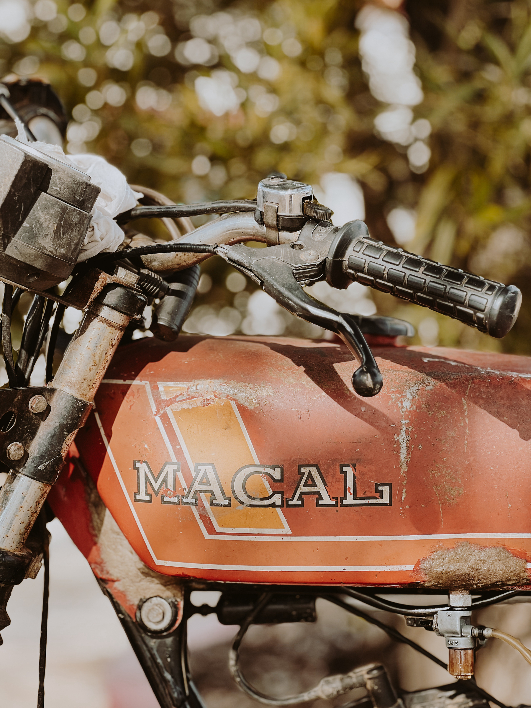Old, weathered motorcycle with the word &ldquo;MACAL&rdquo; on the fuel tank, showing handlebars, throttle grip, and a clutch lever.