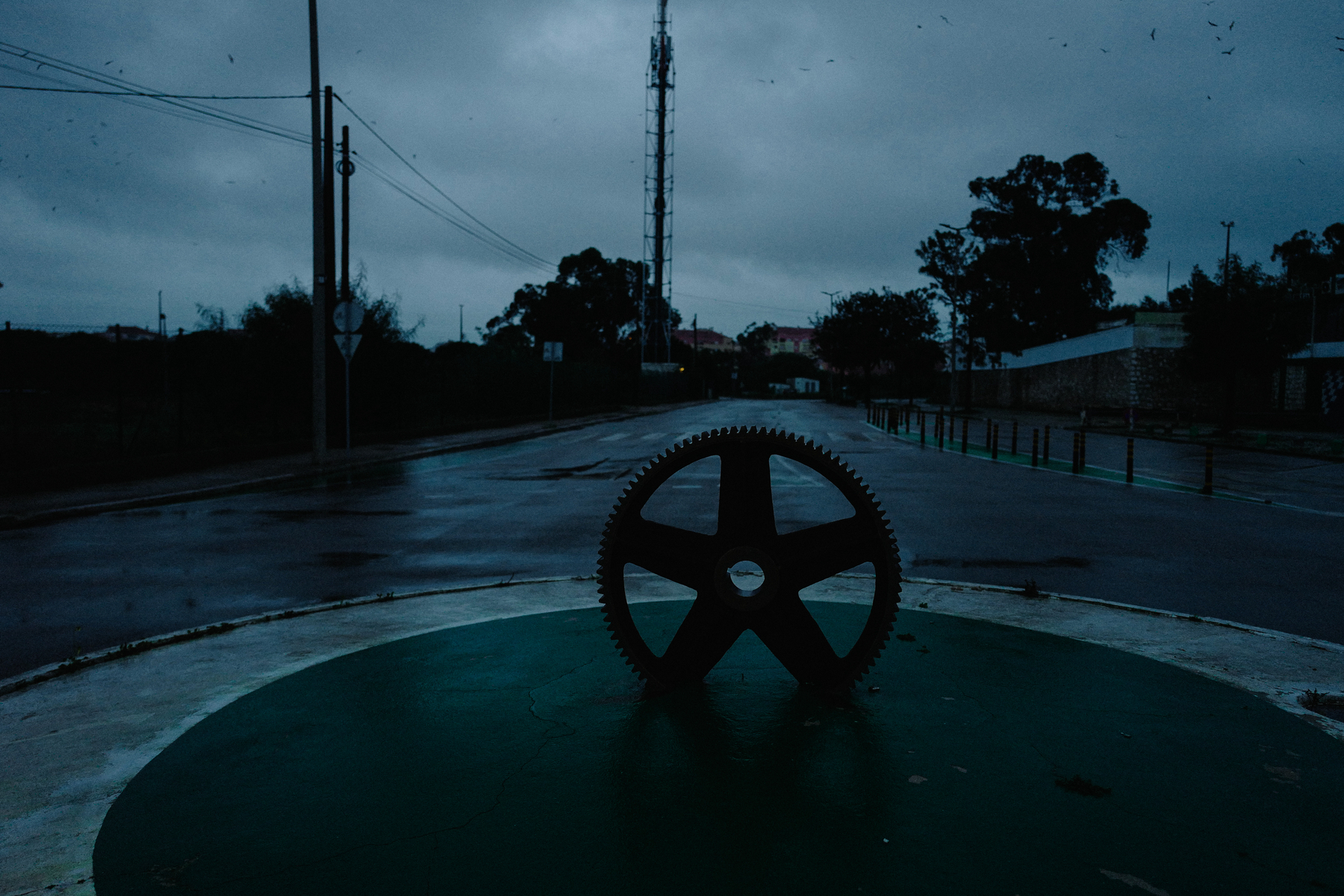 A large gear sculpture on a green circular base in the center of a deserted street at dusk, with a dark cloudy sky and faint silhouettes of birds flying above.