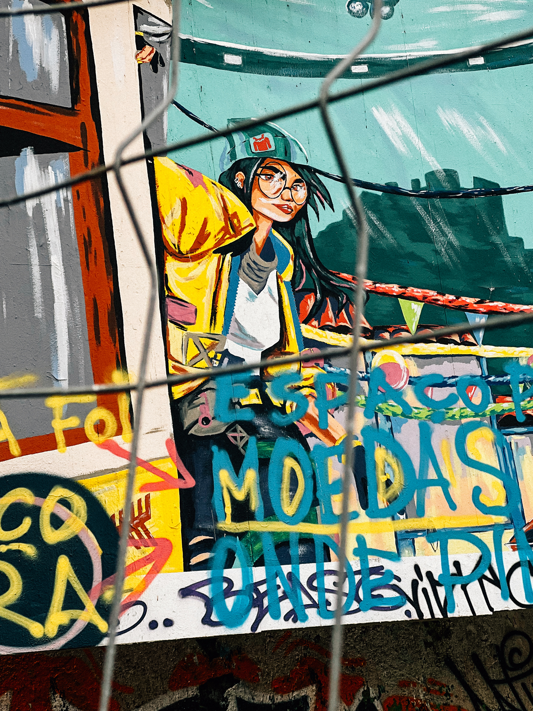 A colorful graffiti mural of a stylish woman with glasses and a hat, seen through a chain-link fence. The foreground features graffiti tags over the art.