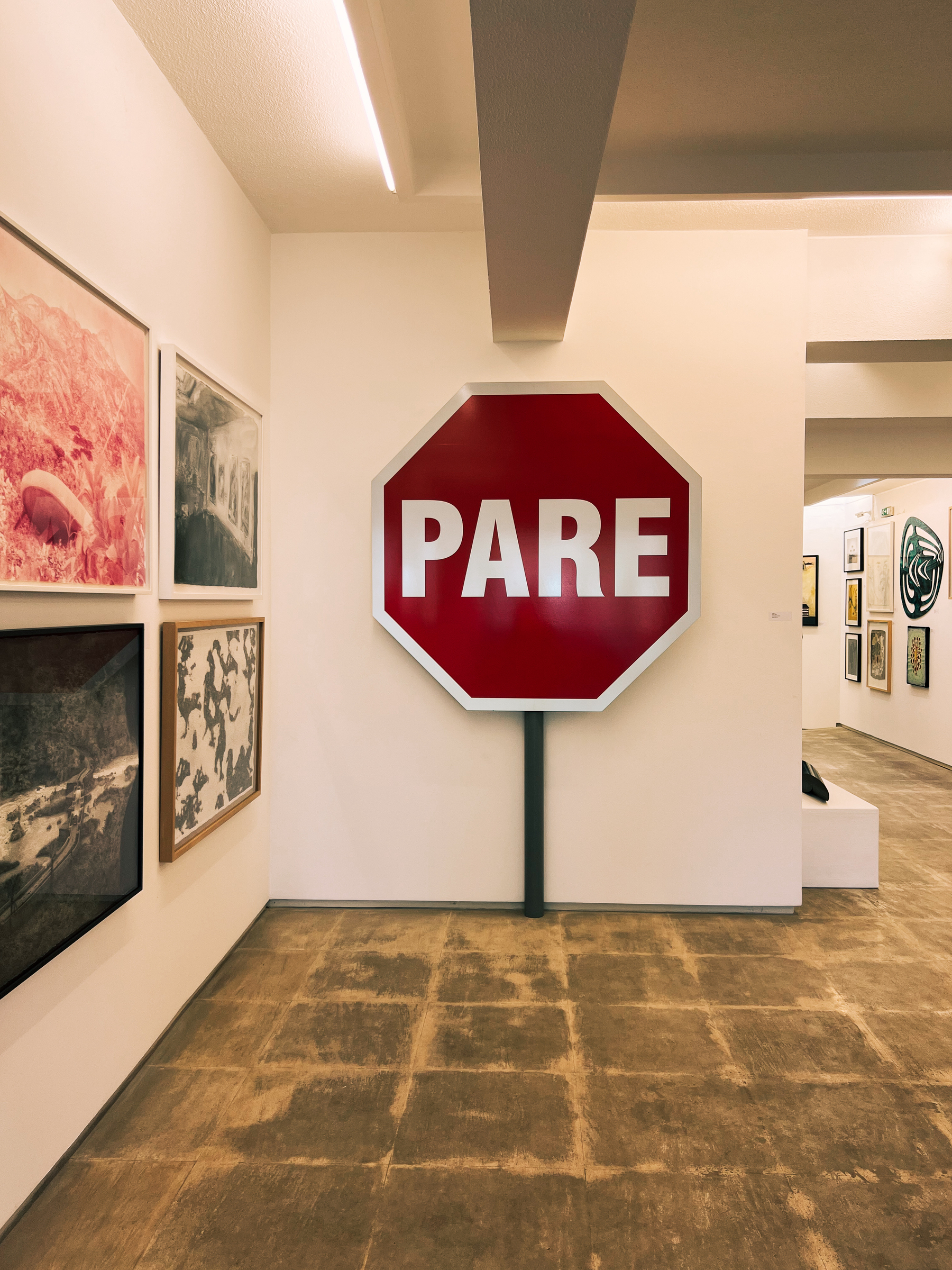 Octagonal stop sign with &ldquo;PARE&rdquo; text displayed in an art gallery with varied artwork on walls and a tiled floor.