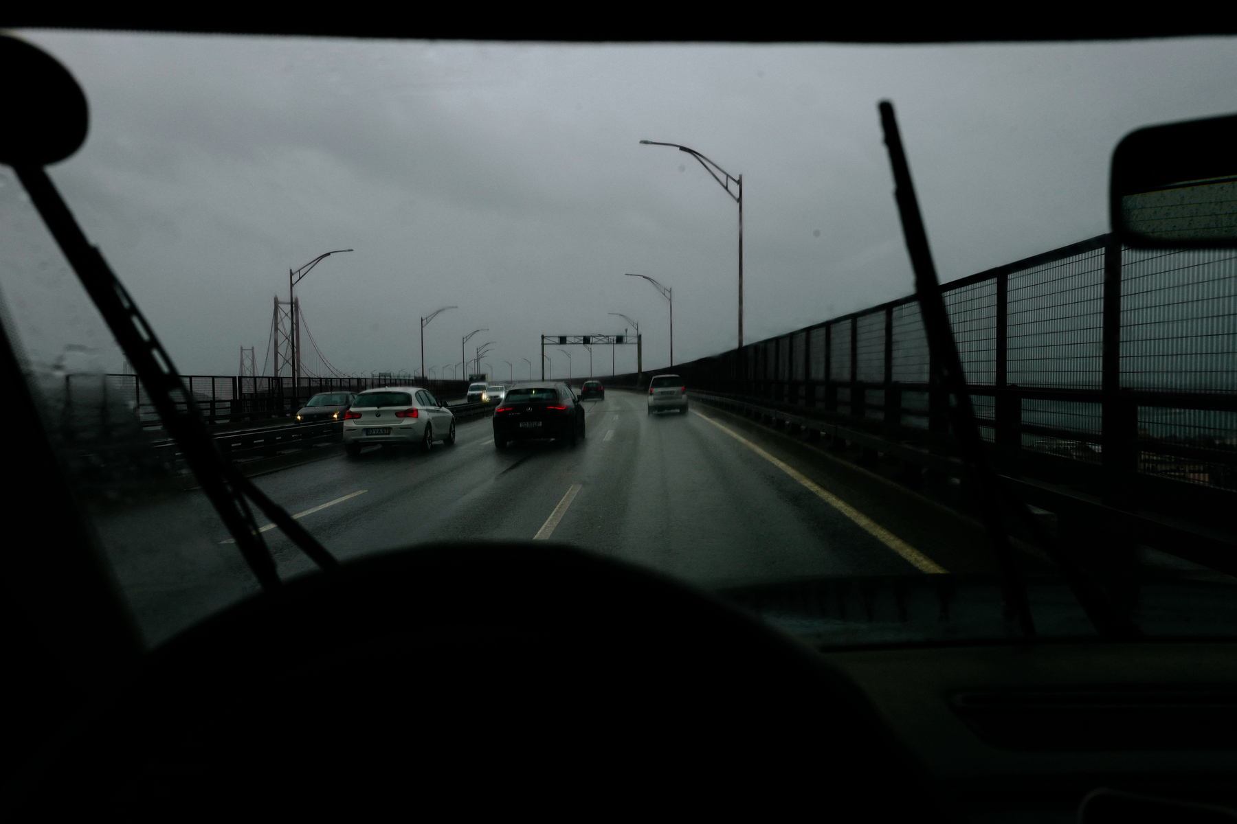 View from inside a vehicle crossing a bridge on a rainy day, with other cars traveling on the road and overcast skies above.