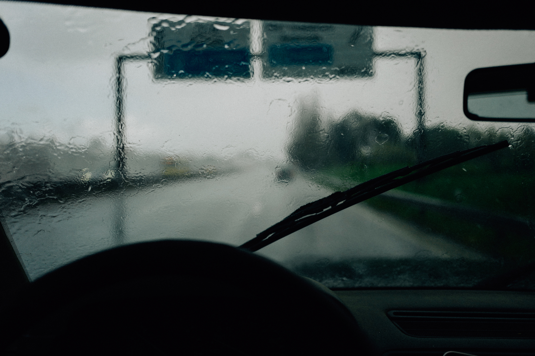 View from inside a car during rain with water droplets on the windshield and windshield wipers visible, looking out onto a blurred road and traffic signs.