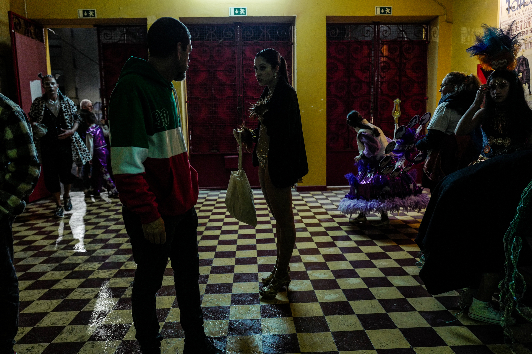 A dimly lit room with a checkered floor where a man and a woman are having a conversation. In the background, individuals dressed in vibrant and elaborate costumes, including feathers, prepare for a performance or festivity.