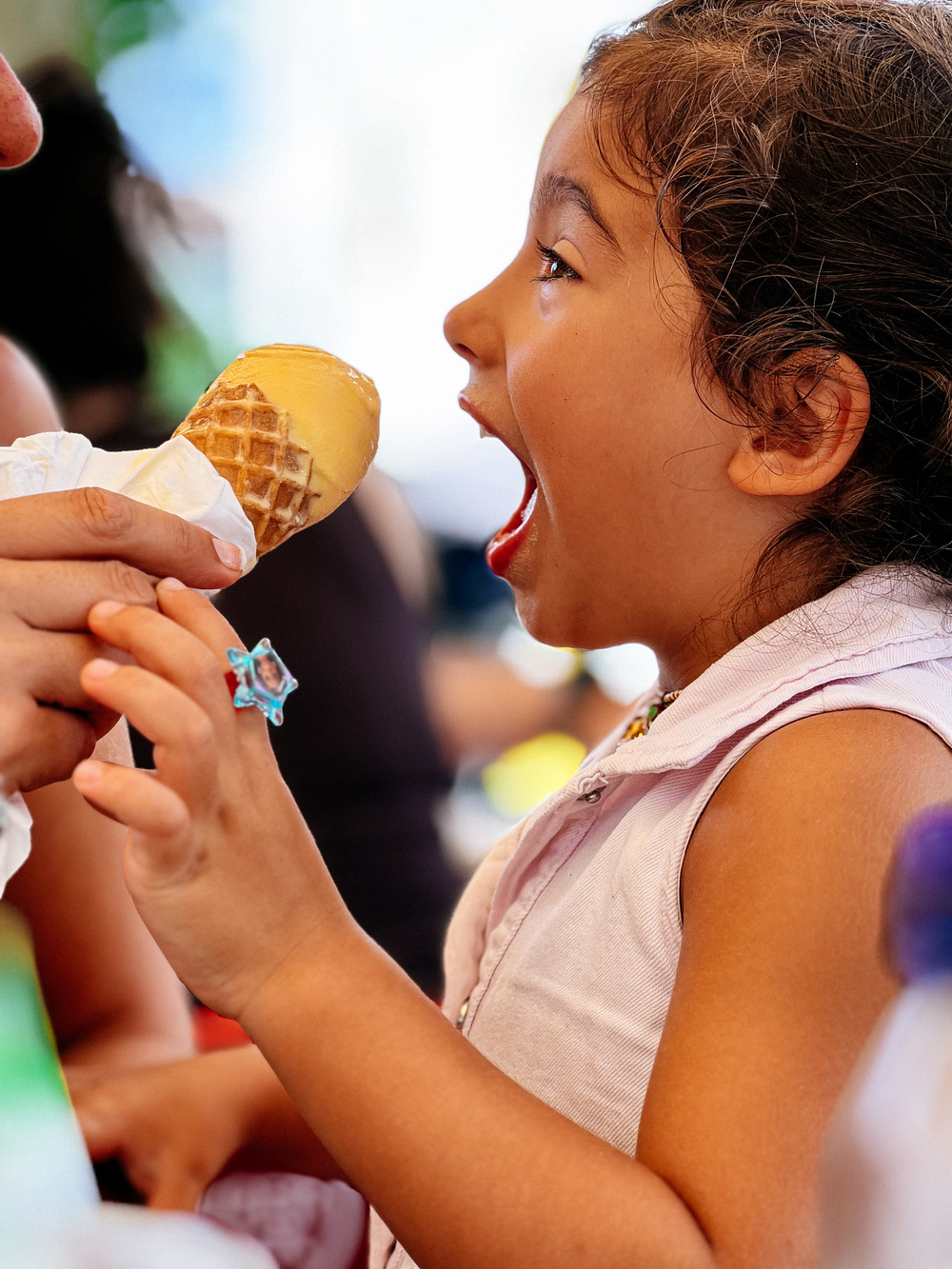 A young girl with an excited expression and open mouth is about to take a bite of an ice cream cone held by an adult. She is wearing a sleeveless top and a colorful ring on her finger.