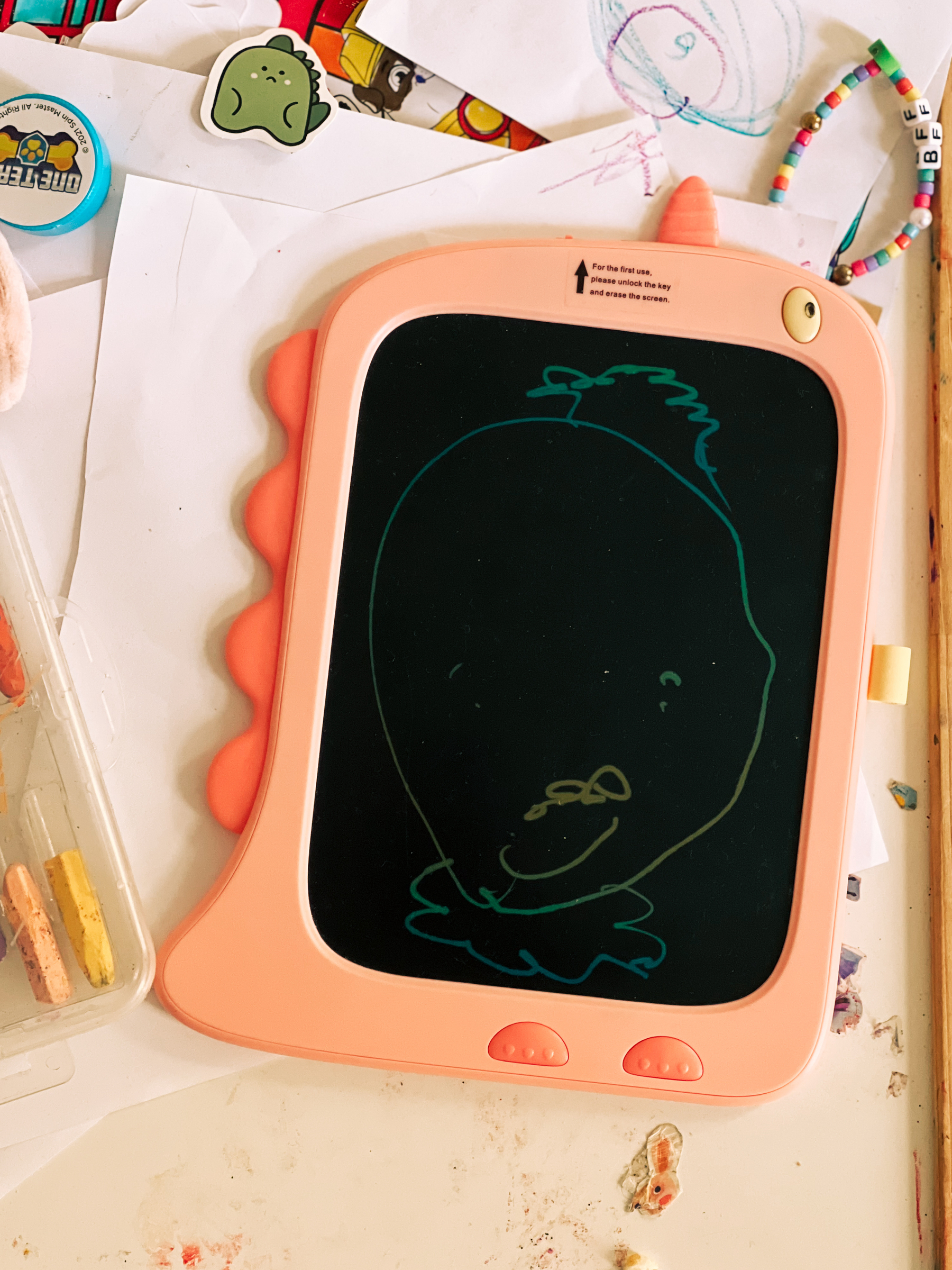 A child&rsquo;s magnetic drawing board with a simple doodle on it, depicting a smiley face with hair, surrounded by various art supplies and drawings on a messy table.