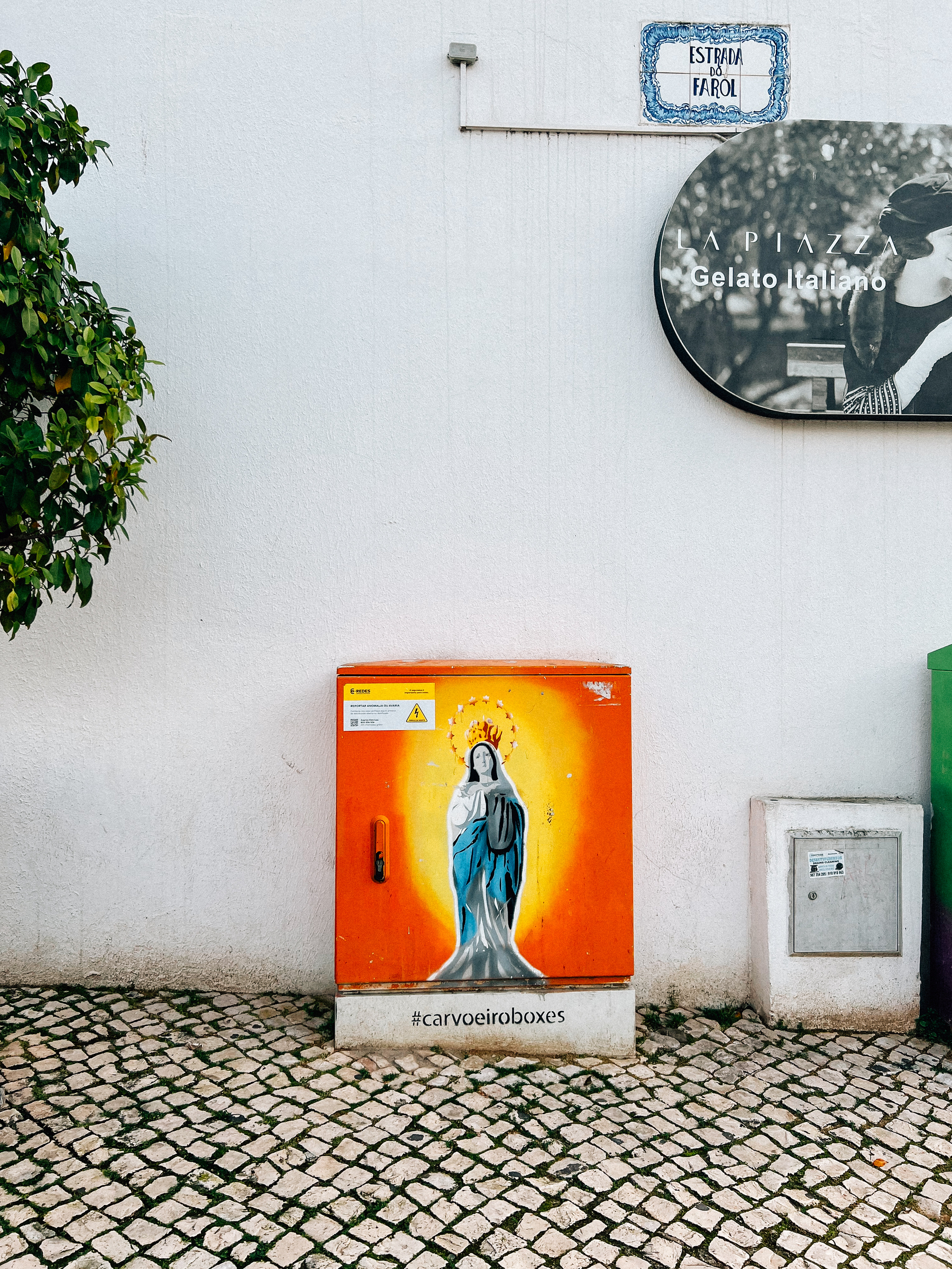 An electrical utility box on a street with a colorful depiction of the Virgin Mary, hashtag #carvoeiroboxes at the bottom, adjacent to a traditional cobblestone pavement and white walls.