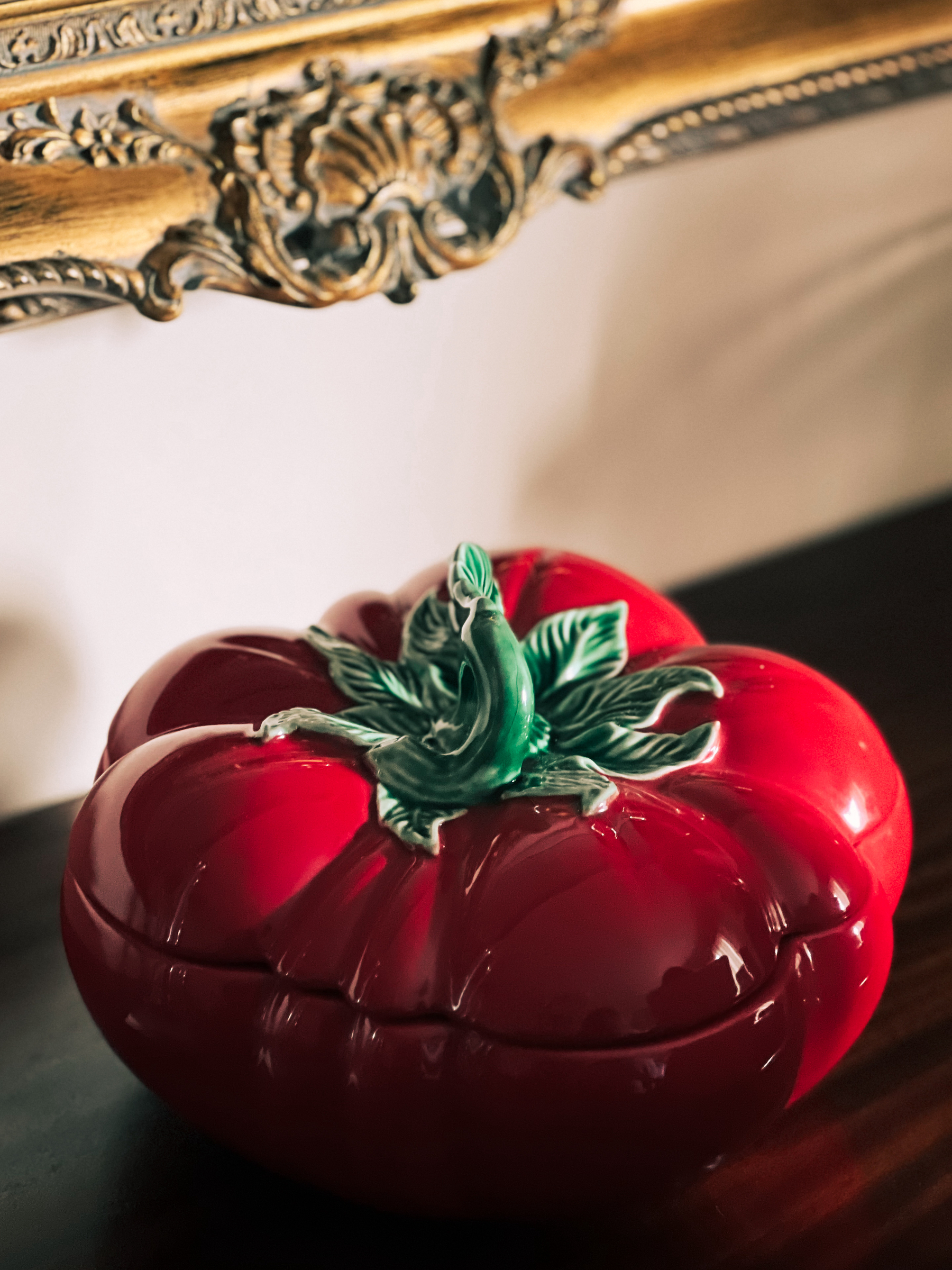 A ceramic container shaped like a red tomato with a green stem sits on a wooden surface against a wall with an ornate gold-framed mirror.