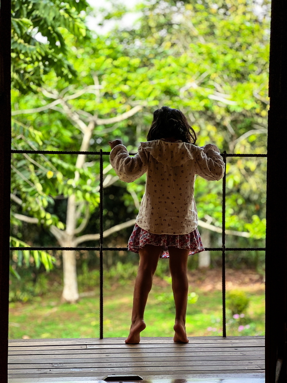 A child is standing barefoot on a wooden deck, holding onto a railing. Lush green trees are visible in the background.