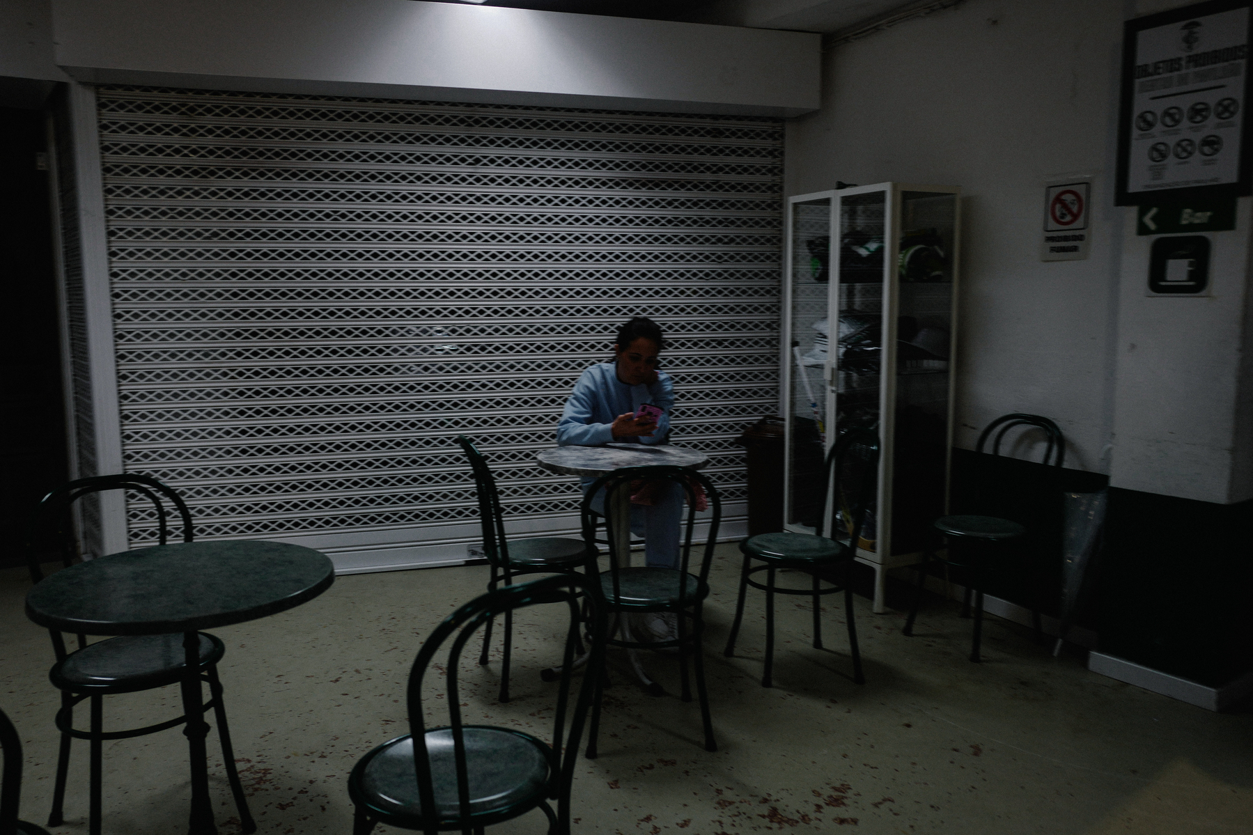 A dimly lit interior of a café with a closed metal security gate. In the center, a person sits at a small round table, engrossed in their smartphone. There are empty chairs and another table nearby, with a display cabinet and a