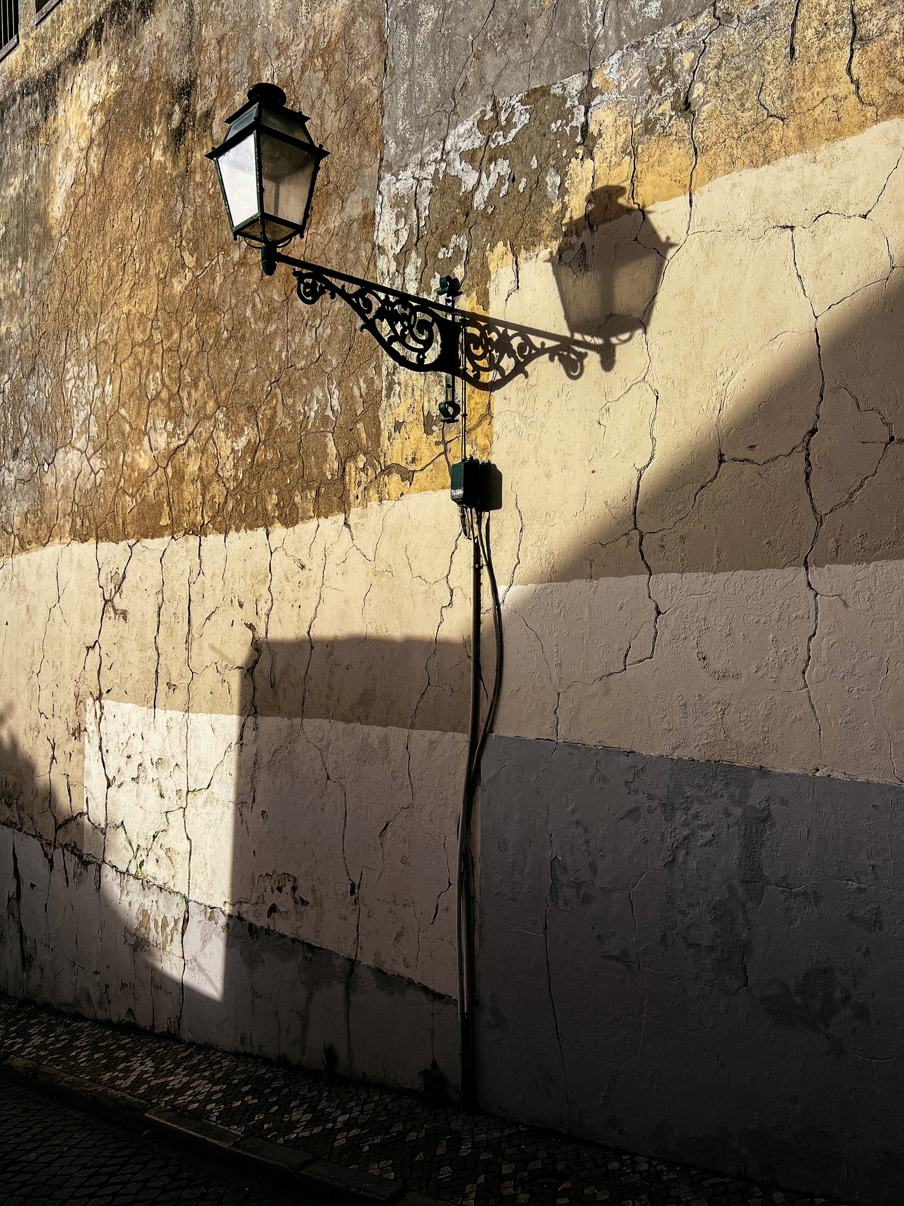 Street lamp mounted on a decorative metal bracket projecting from a weathered wall, casting a shadow, with cobbled street below.