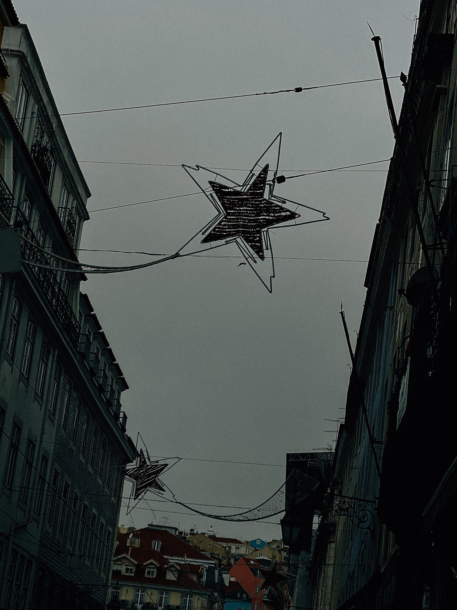Street view with decorative star-shaped lights strung across wires above, between buildings, under an overcast sky.