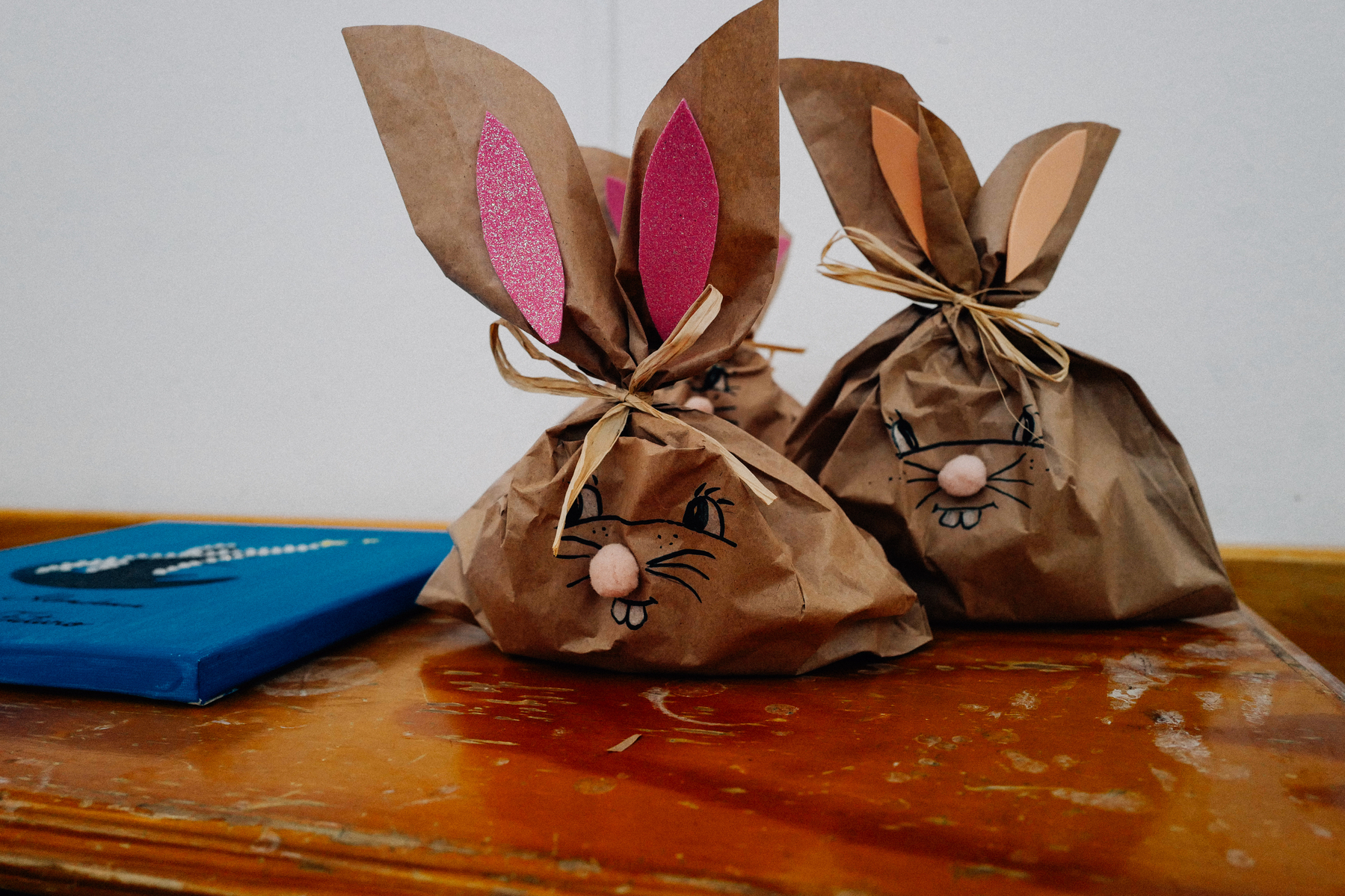 Three bunny-shaped gift bags with drawn faces and ears made of paper on a wooden table, with a book lying to the side.