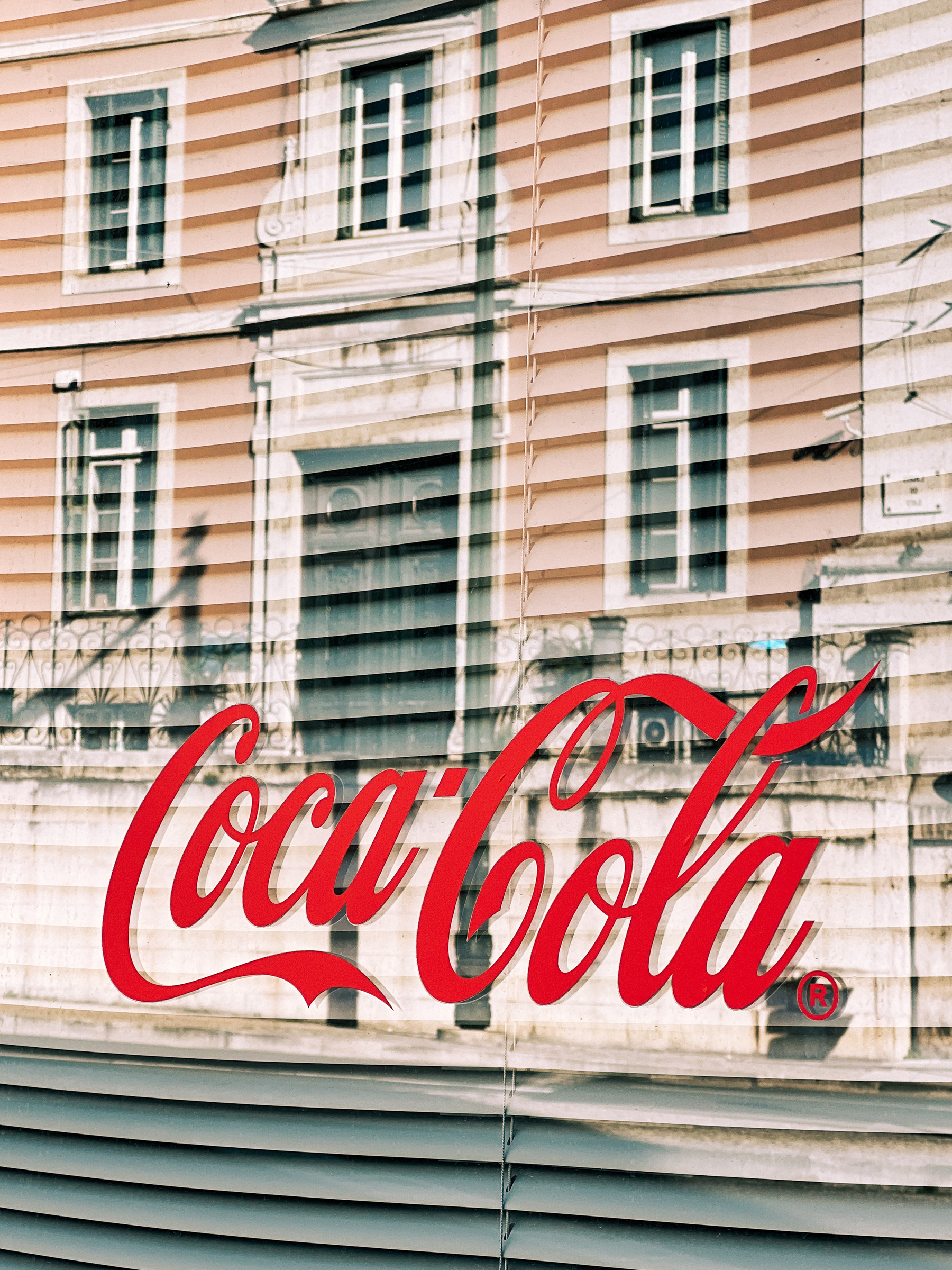 A Coca-Cola logo in red script overlaid on the reflection of a striped building facade in a window.