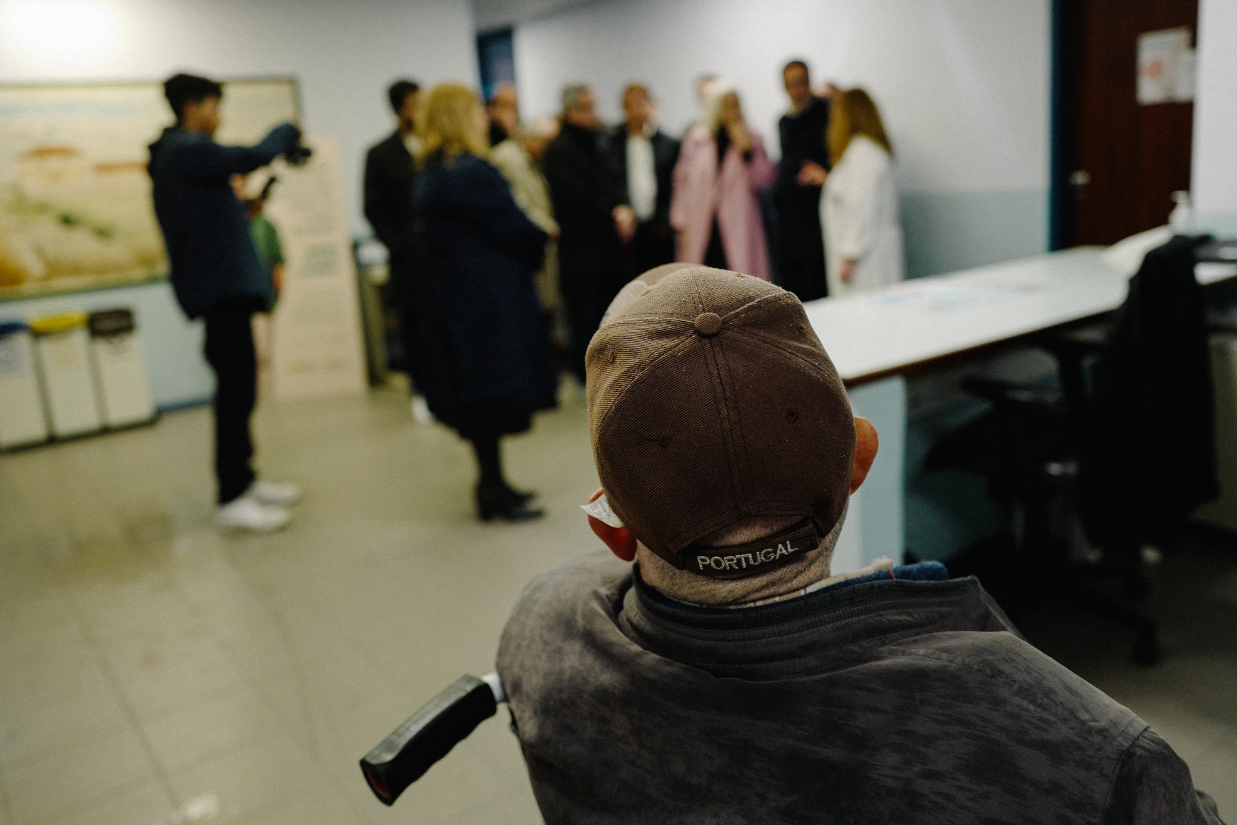 A person in a wheelchair wearing a hat with &ldquo;Portugal&rdquo; on it is facing a group of standing people who appear to be engaged in a conversation, while another person is taking a photo of the group. The setting seems to be an indoor room.