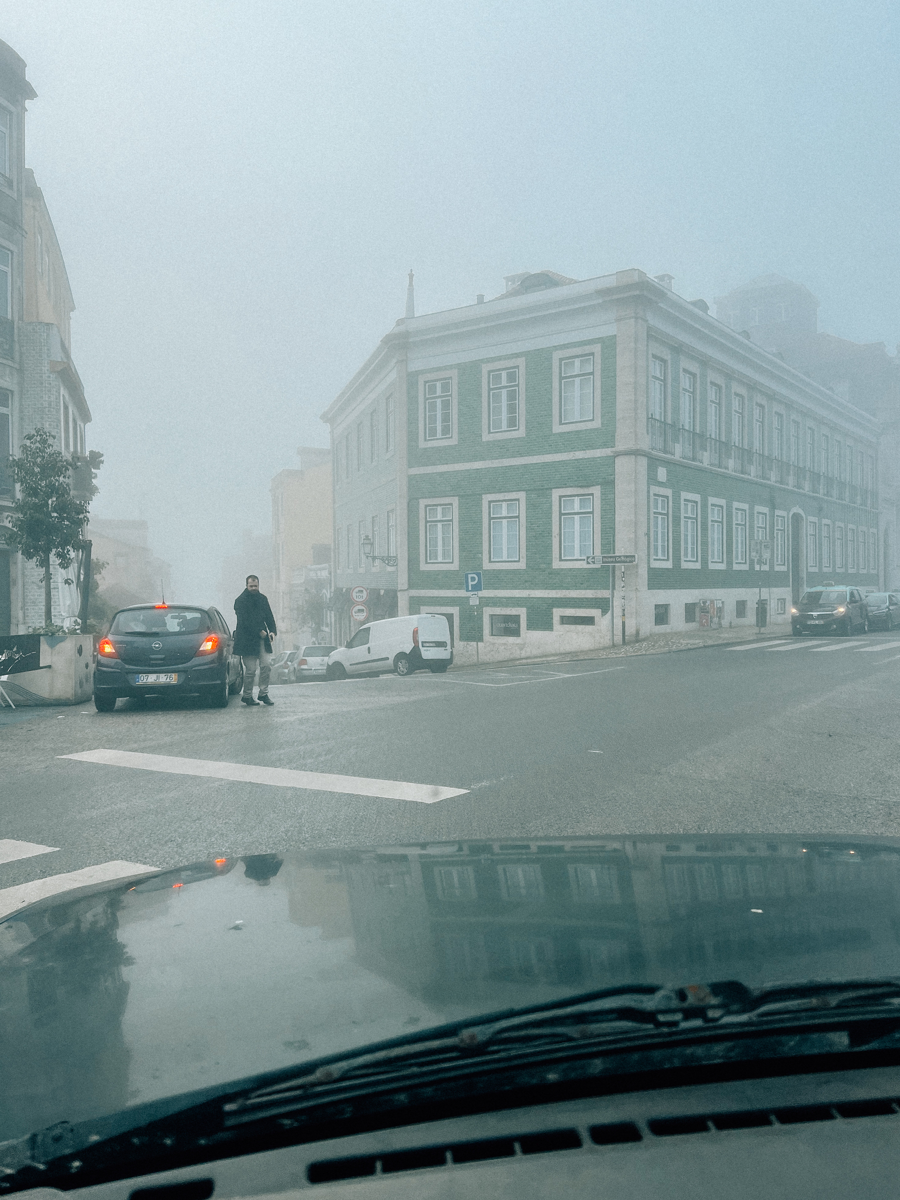 Foggy city street scene with a pedestrian crossing the road, vehicles parked on the side, and a green building on the corner, viewed from inside a car through the windshield.