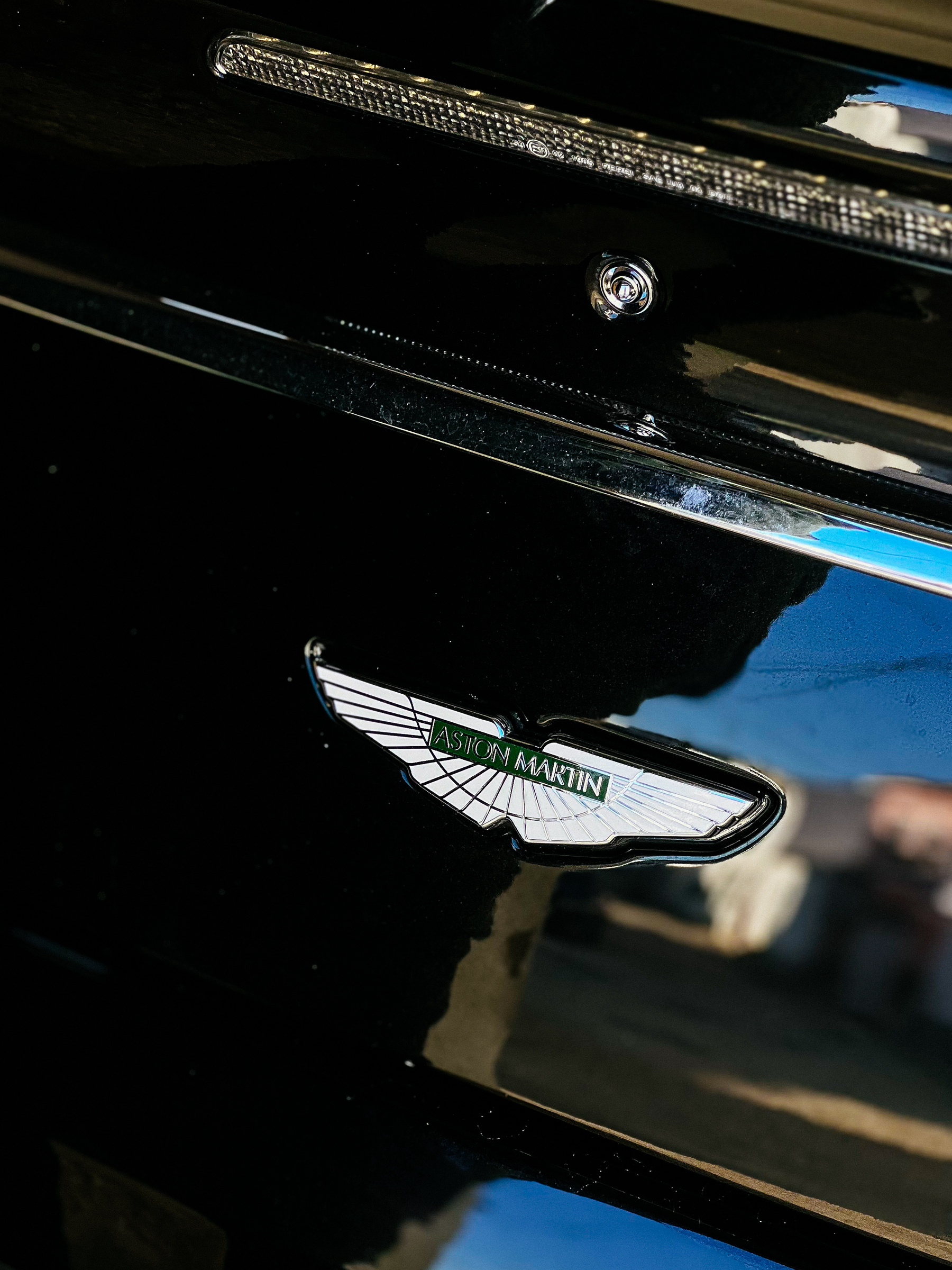 An Aston Martin badge on a glossy black car surface, with reflections visible around the emblem.