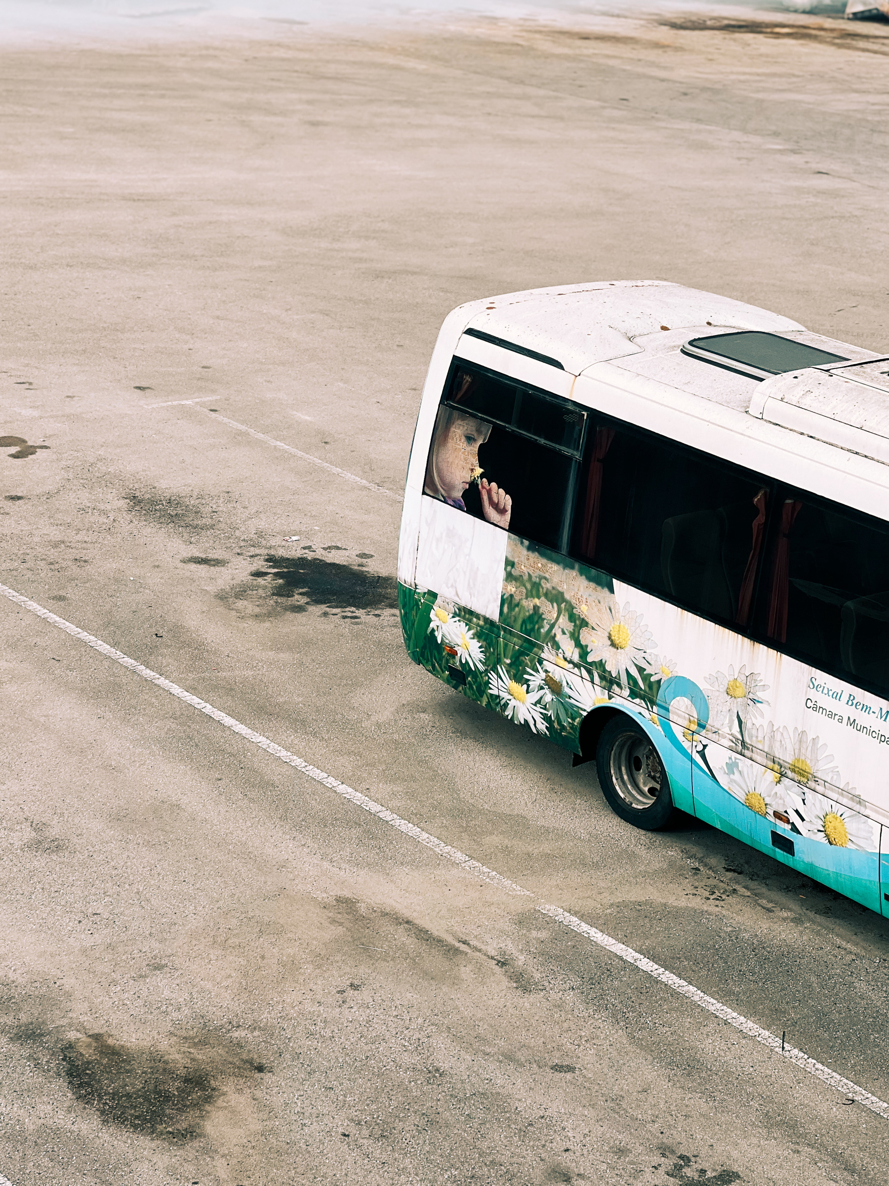 A person looking out from a partially visible bus with a floral design, parked on an expanse of tarmac with visible road markings.