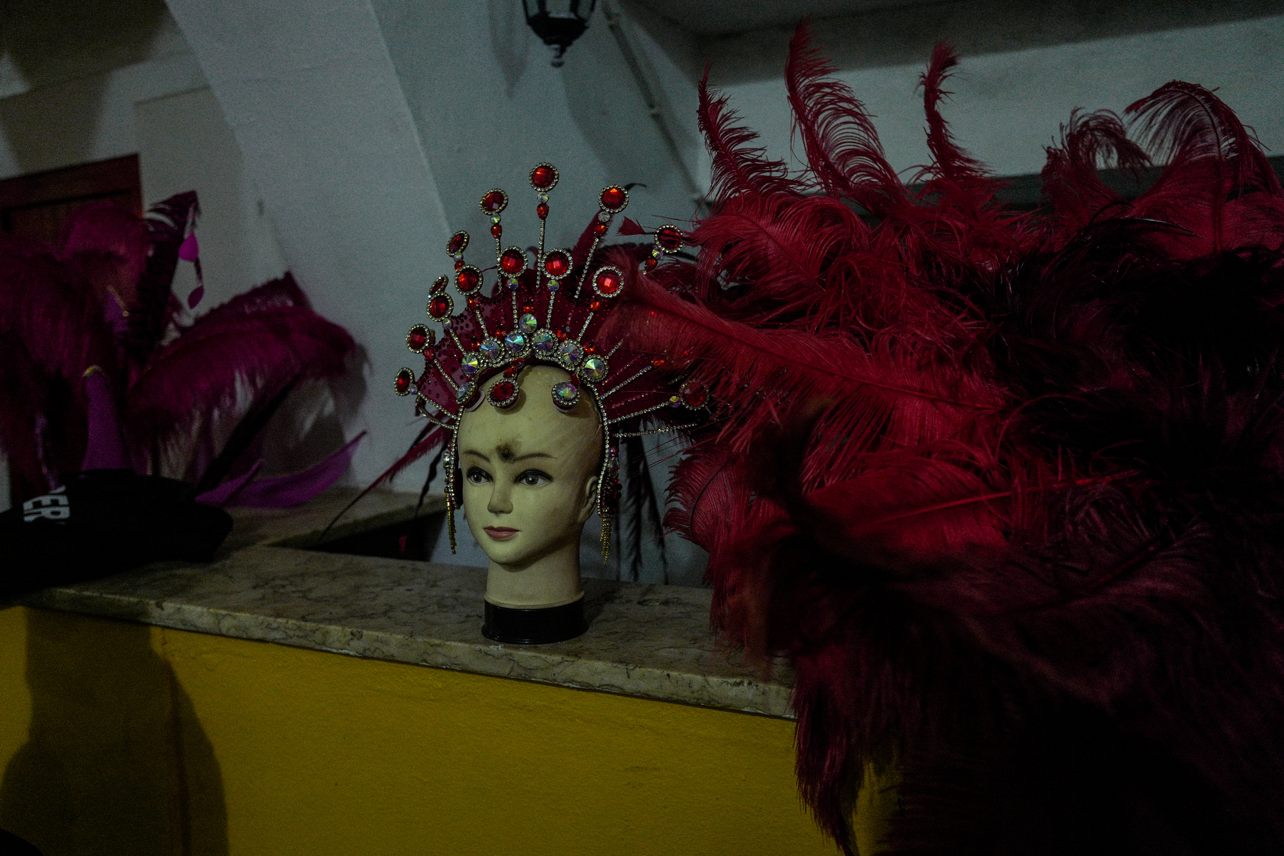 A mannequin head wearing a traditional Chinese headdress adorned with red feathers and gemstones, positioned on a shelf next to red feathered accessories.