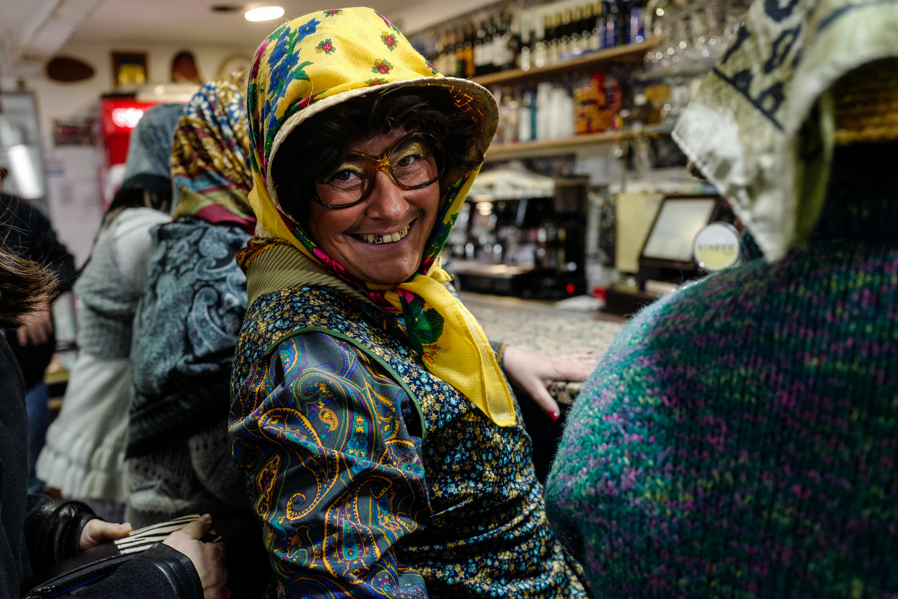 A smiling person wearing glasses, a patterned headscarf, and a colorful jacket inside a bar or café. Other patrons are in the background.