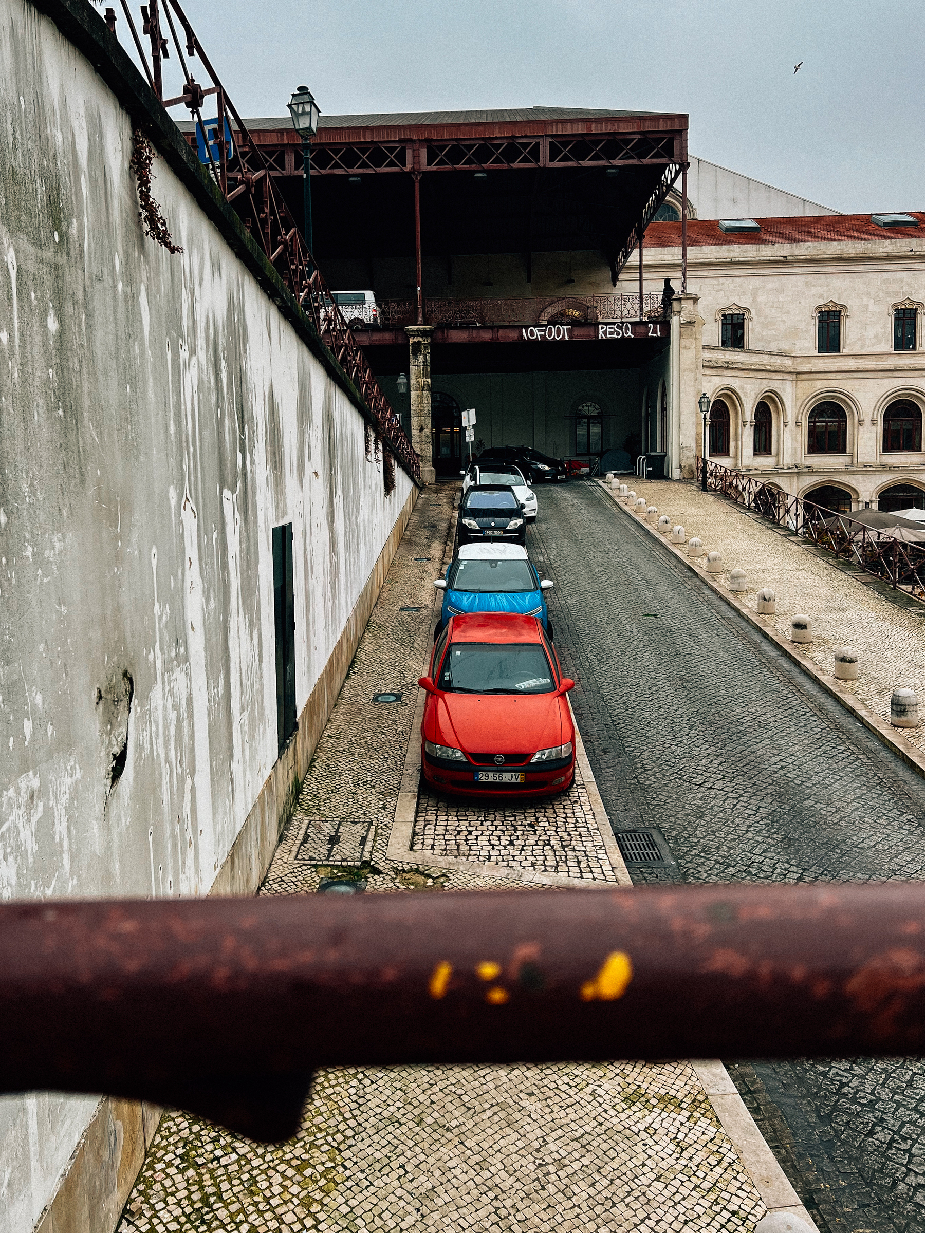 A narrow alley with a line of parked cars, framed by a weathered wall on the left and an industrial building with a red iron bridge structure to the right.