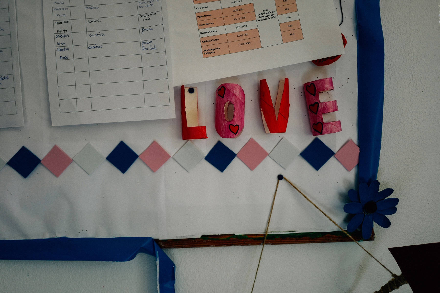 A wall with decorative elements and papers. The word &ldquo;LOVE&rdquo; in colorful painted letters, a blue paper flower, diamond-shaped paper cutouts in pink and blue, and charts with handwritten text.