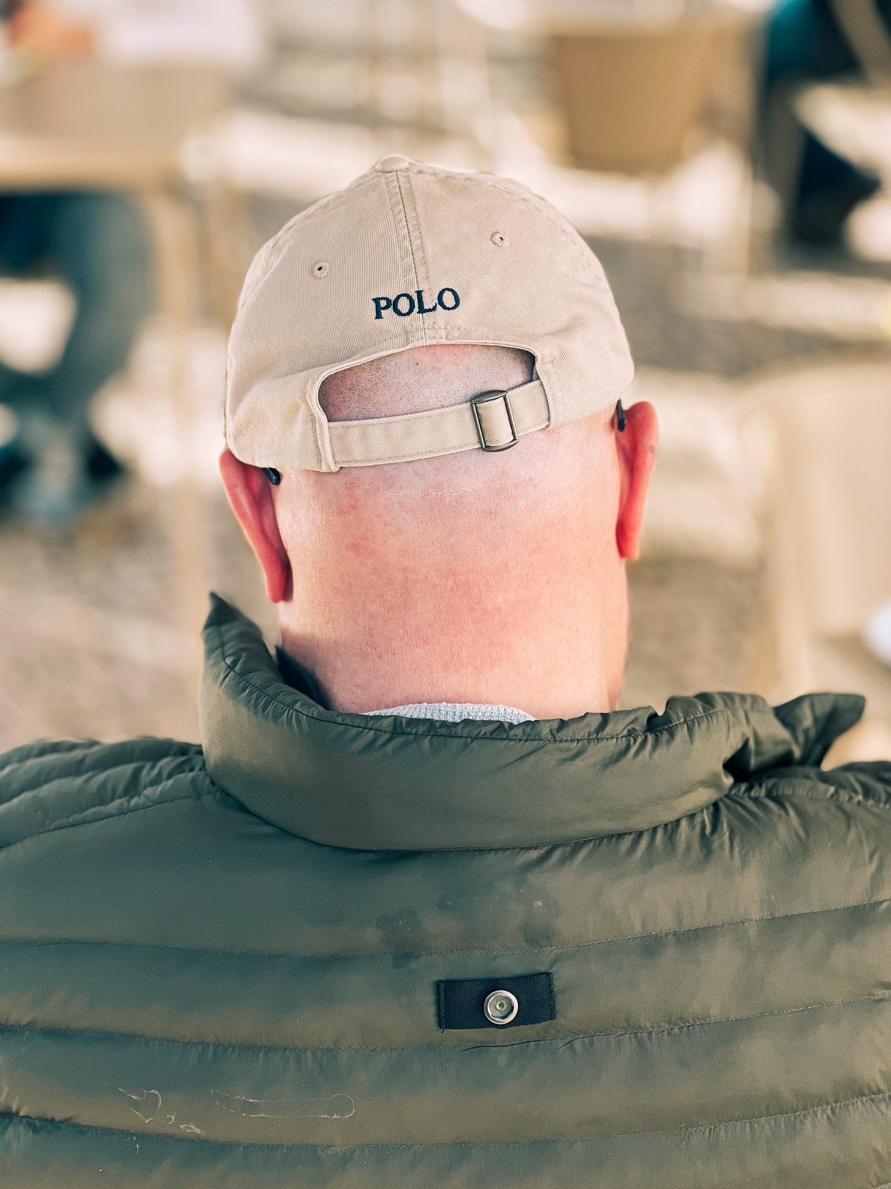 A person from behind wearing a beige baseball cap and a green quilted jacket. The person&rsquo;s neck shows signs of sunburn.
