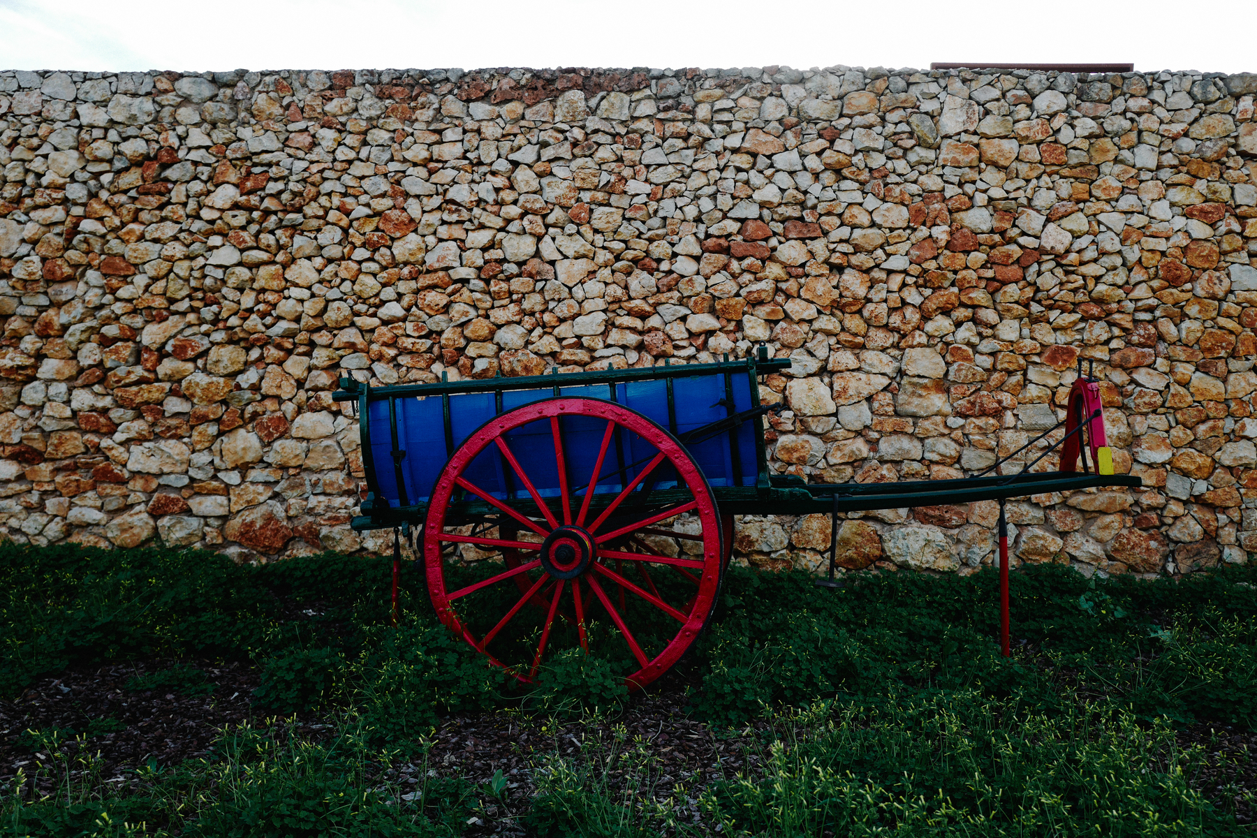 A colorful wooden cart with red wheels is parked in front of a stone wall.