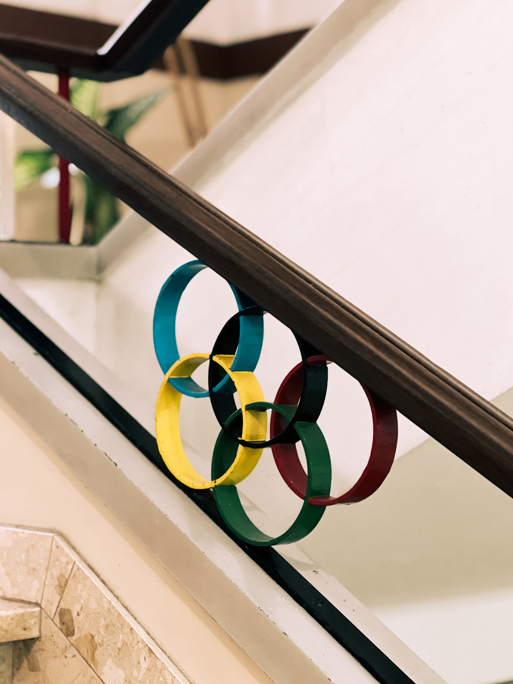 Five interlocking rings in blue, yellow, black, green, and red, representing the Olympic rings, are mounted on a stairway railing with a staircase in the background.