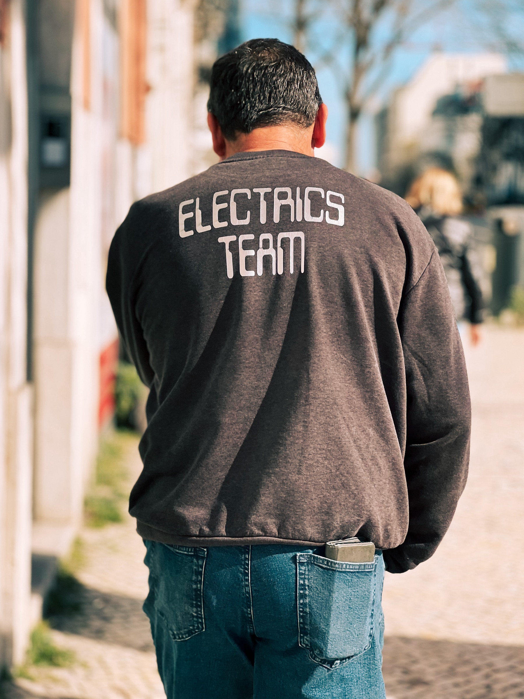 A man standing outdoors wearing jeans and a sweatshirt with the text &ldquo;ELECTRICS TEAM&rdquo; on the back.