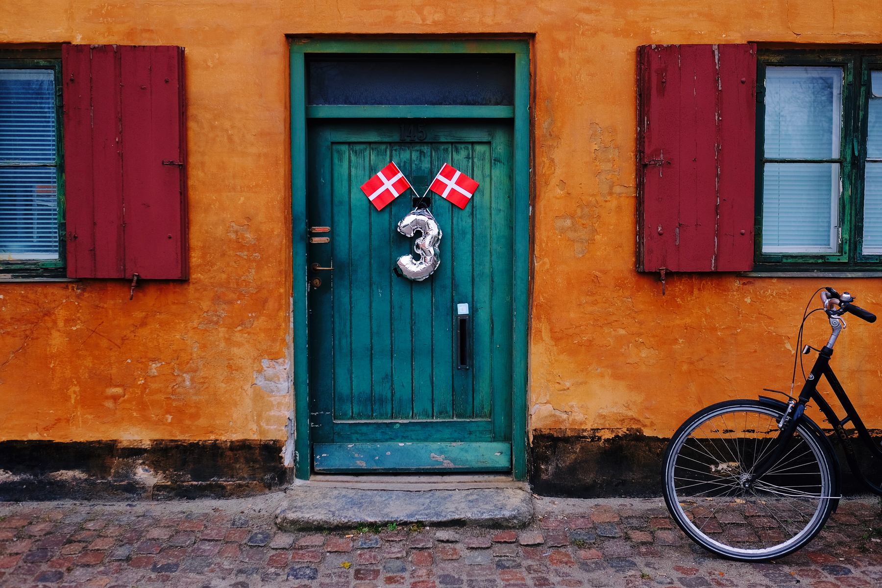 A colorful scene with an orange wall, a teal wooden door flanked by red shutters, Danish flags, a silver balloon shaped like the number &ldquo;3&rdquo;, and a parked bicycle to the right.