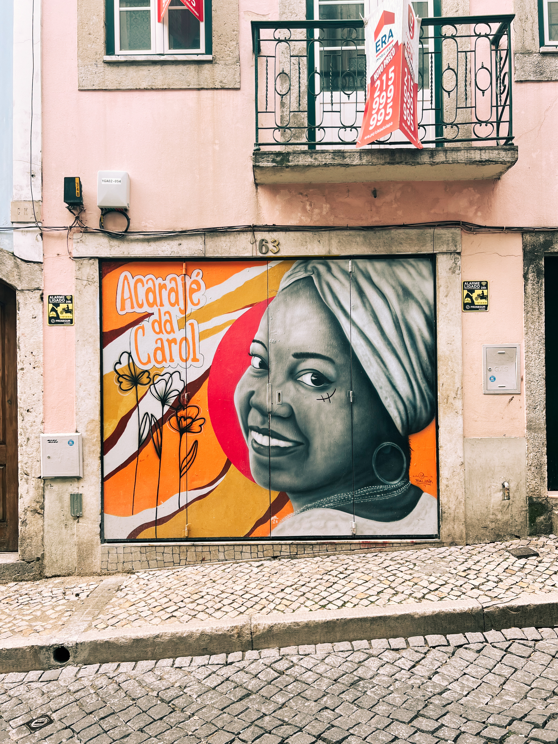 An urban scene showing a colorful street mural of a woman&rsquo;s smiling face on a building, with &ldquo;Acarajé da Carol&rdquo; written beside it. Above the mural, a balcony with a &ldquo;For Sale&rdquo; sign. Cobblestone street.
