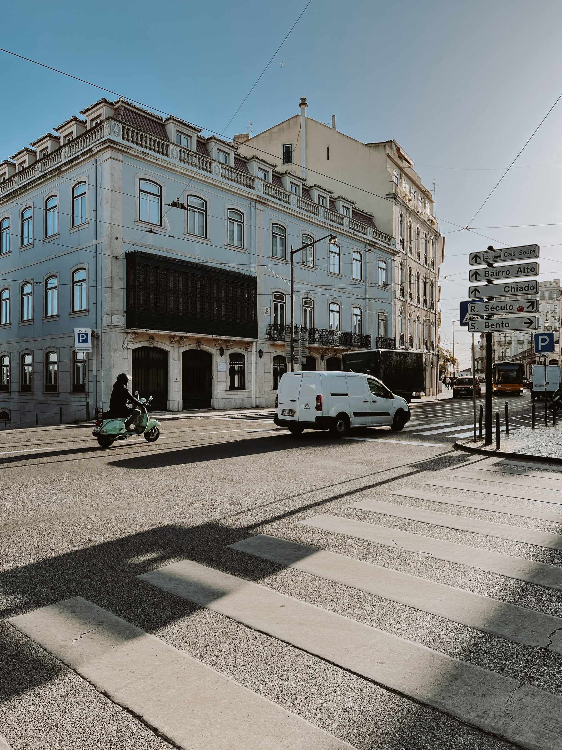 A sunny street scene with traditional European architecture, a pedestrian crossing, a scooter, a white van, and street signs.