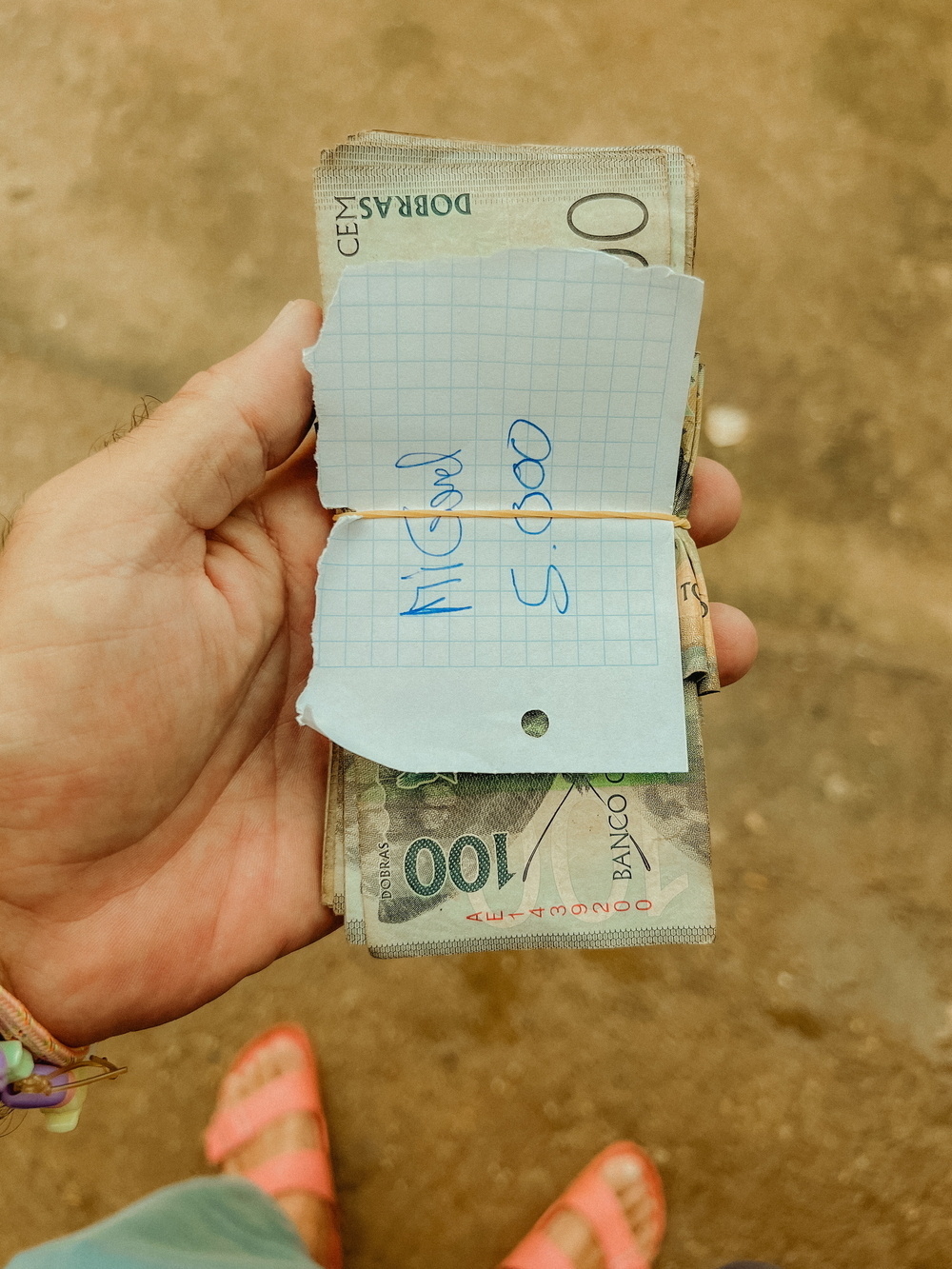 A hand holding a stack of banknotes, secured with an elastic band. On top of the stack is a piece of graph paper with handwritten text that reads &ldquo;Miguel 5.000.&quot;