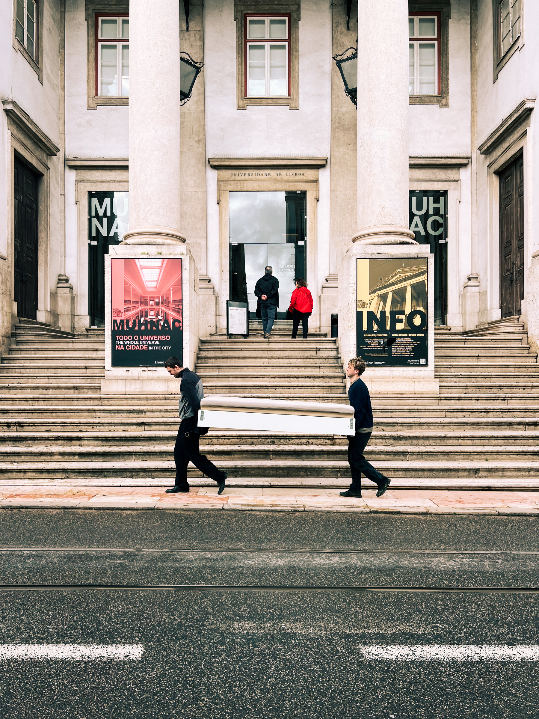 Two individuals carrying a long object on a city street in front of a building with steps and columns, a museum or university entrance, with posters on either side of the doorway.