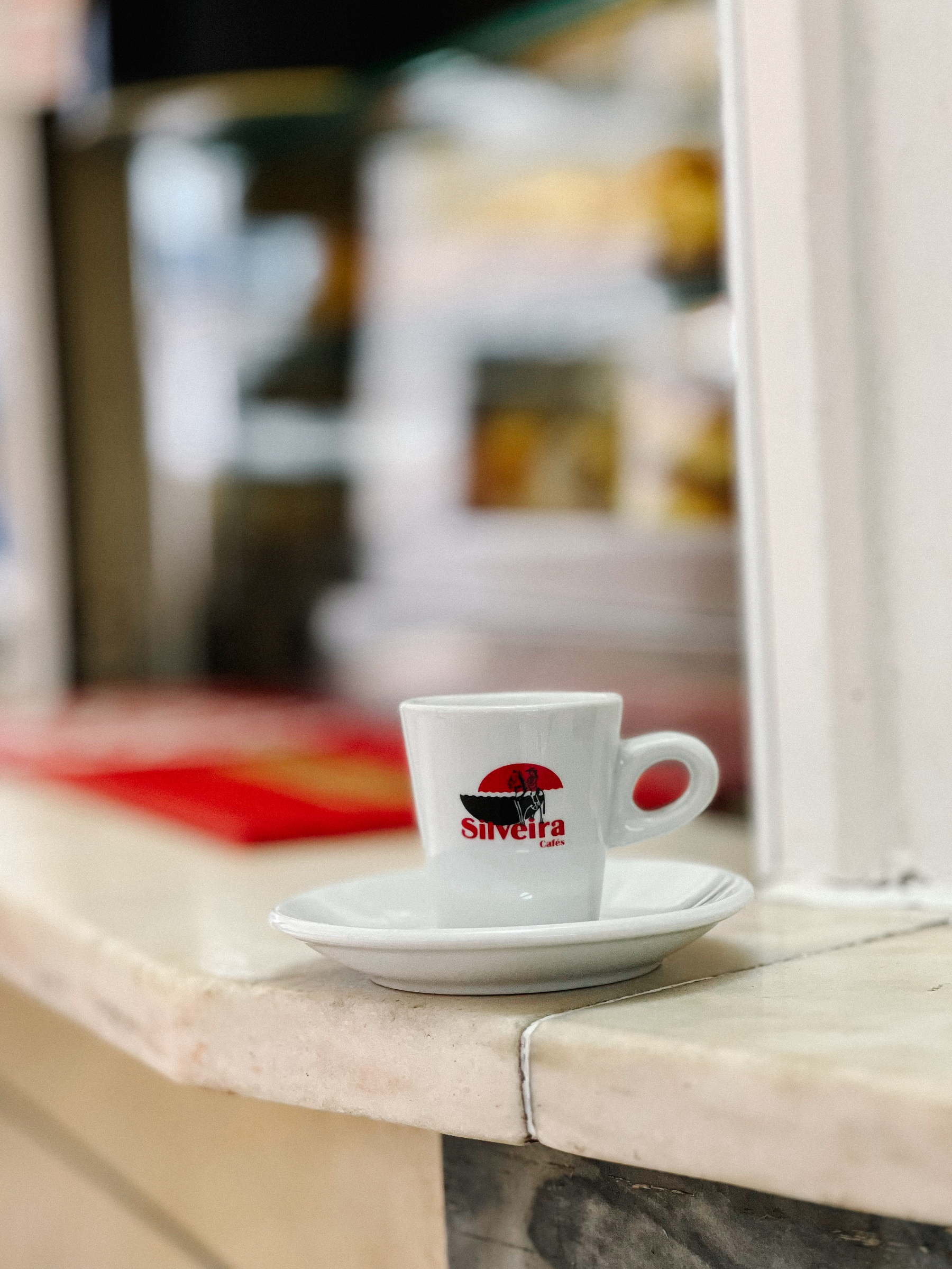 A white espresso cup with &ldquo;Silveira Cafes&rdquo; printed on it, sitting on a matching saucer, placed on the edge of a windowsill. The background is softly blurred.