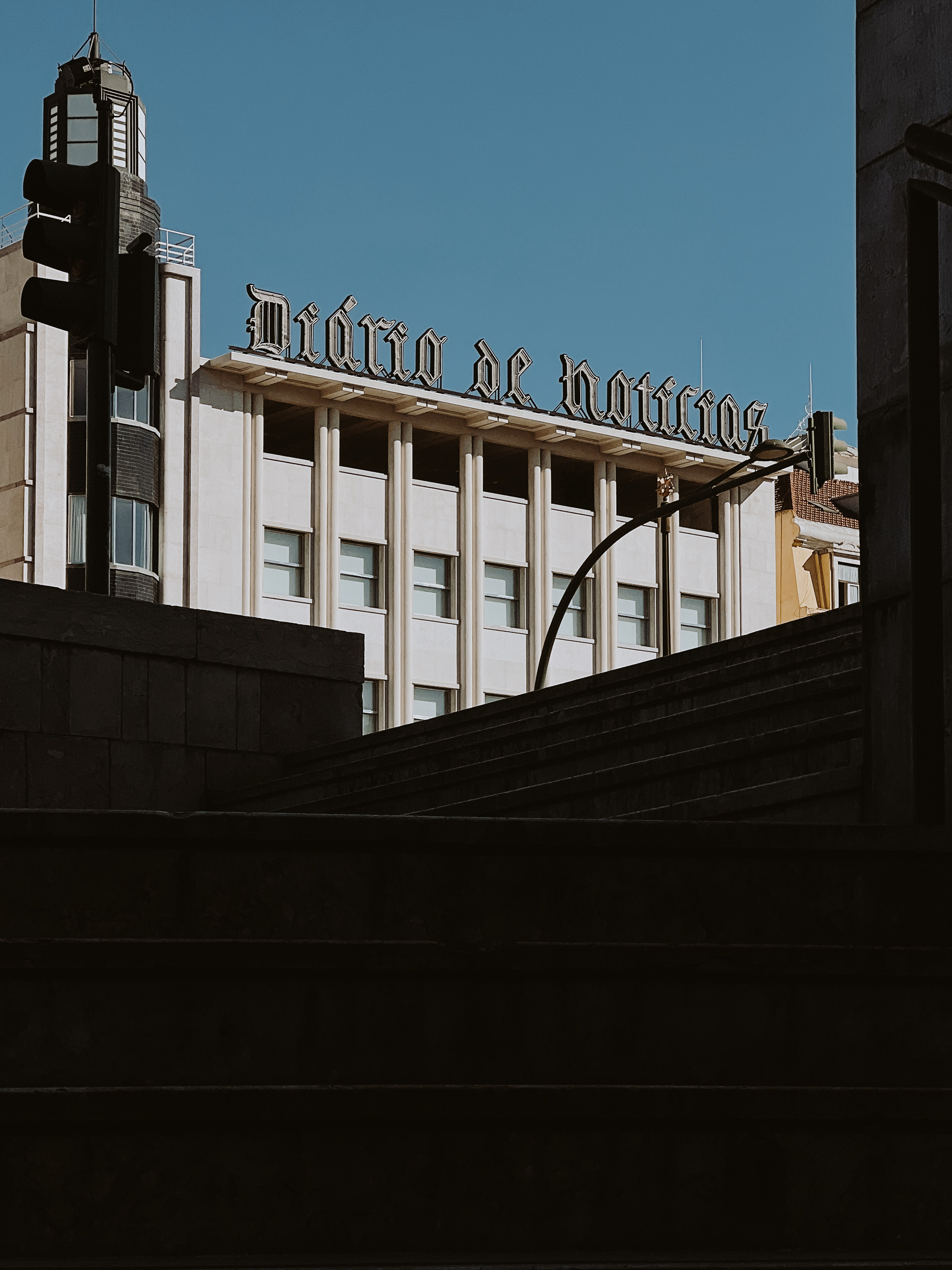 A building with the sign &ldquo;Diário de Notícias&rdquo; atop, visible over a flight of stone steps, with a streetlamp and traffic lights in the foreground against a clear sky.