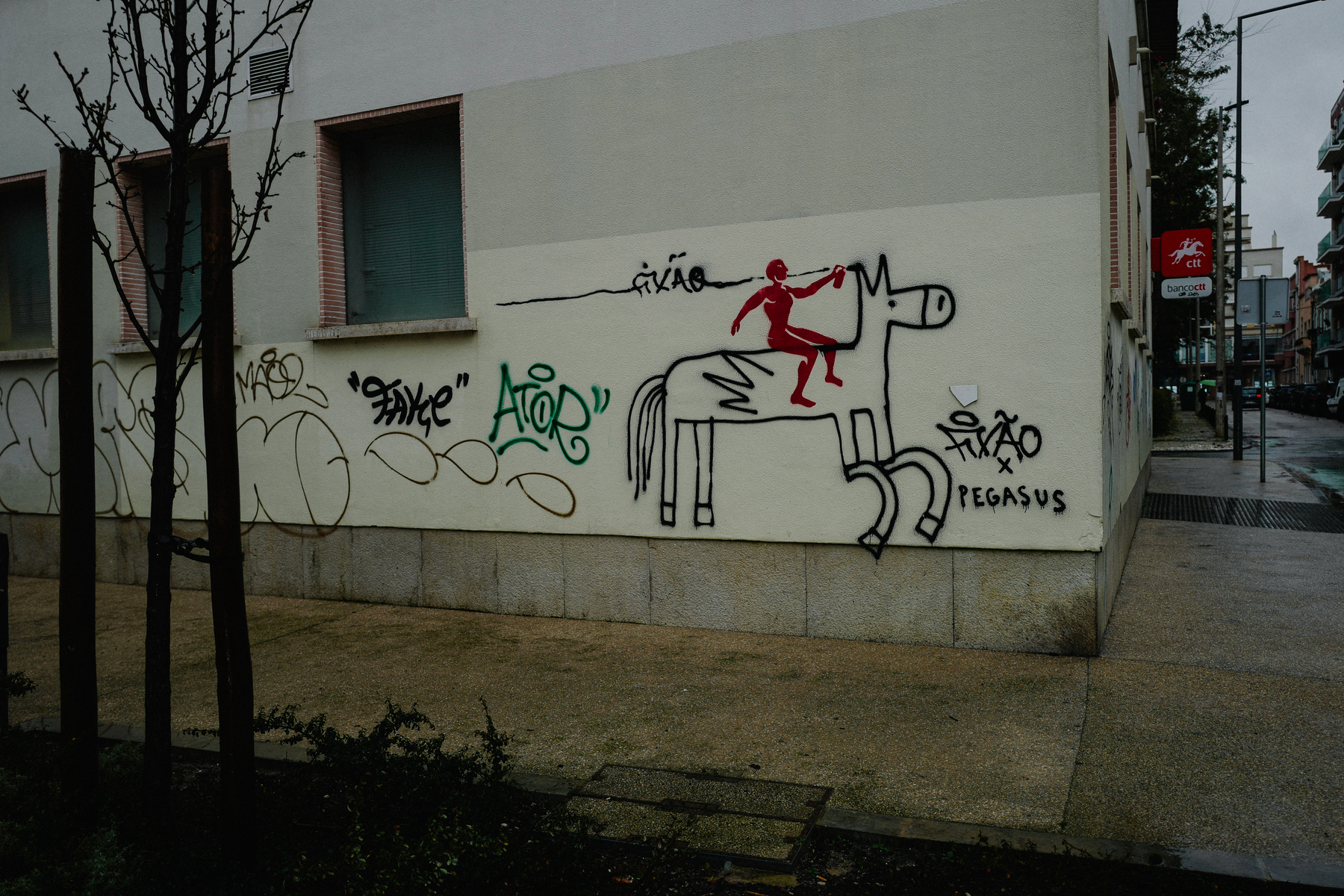 A street wall with graffiti depicting a stick figure human riding a simplistic horse, surrounded by various graffiti tags and writings. There&rsquo;s a bare tree in the foreground and a street scene with buildings in the background.