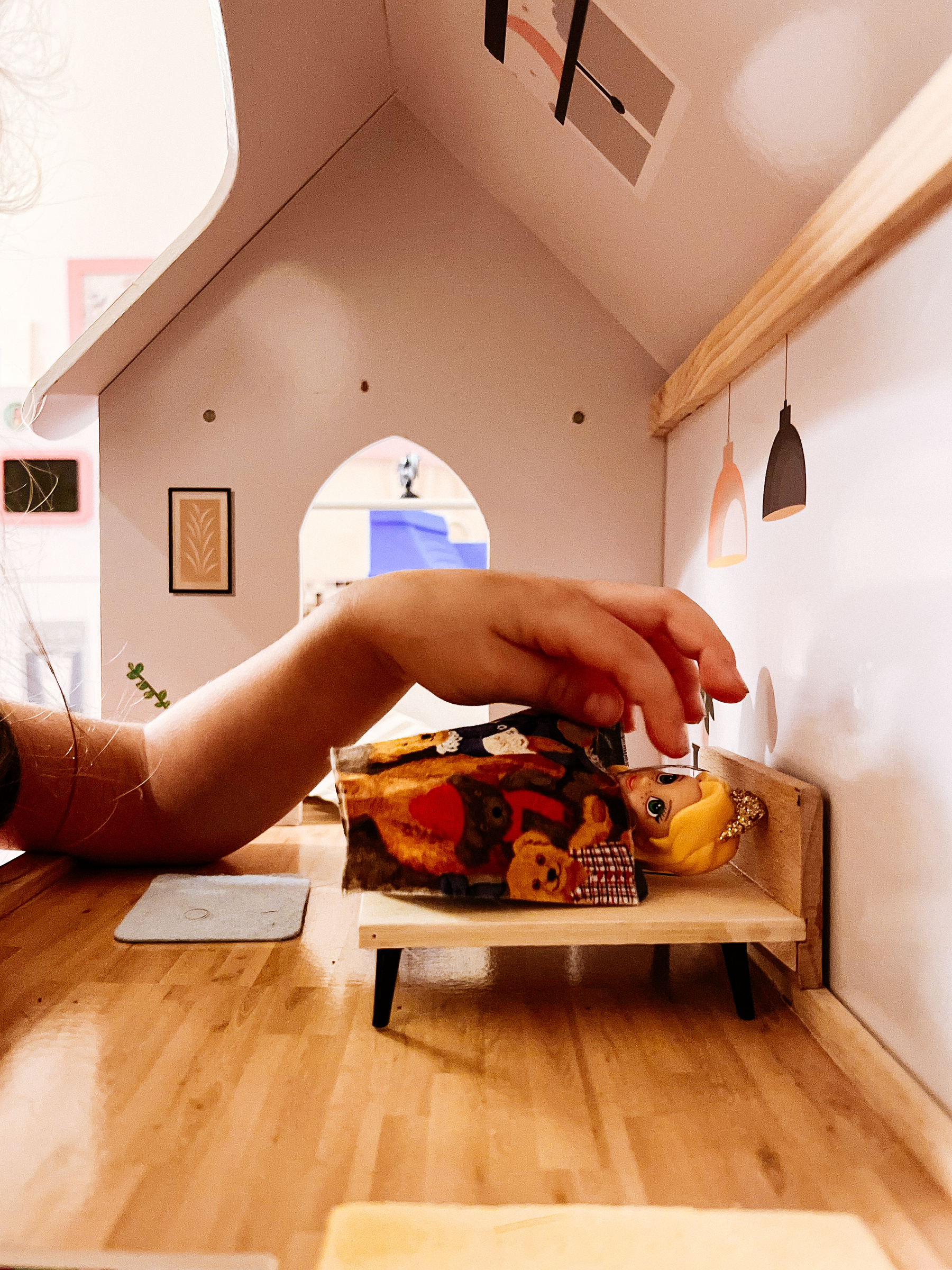 A child&rsquo;s hand playing with a small toy figure inside a dollhouse, focusing on arranging the figure on a miniature bed.