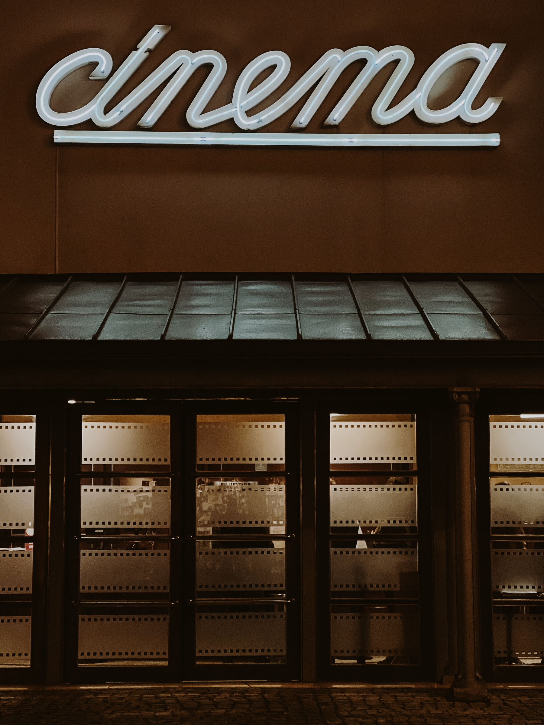 A nighttime view of a cinema entrance with a neon sign above the door. The facade is warmly lit, highlighting the sign and the front doors.