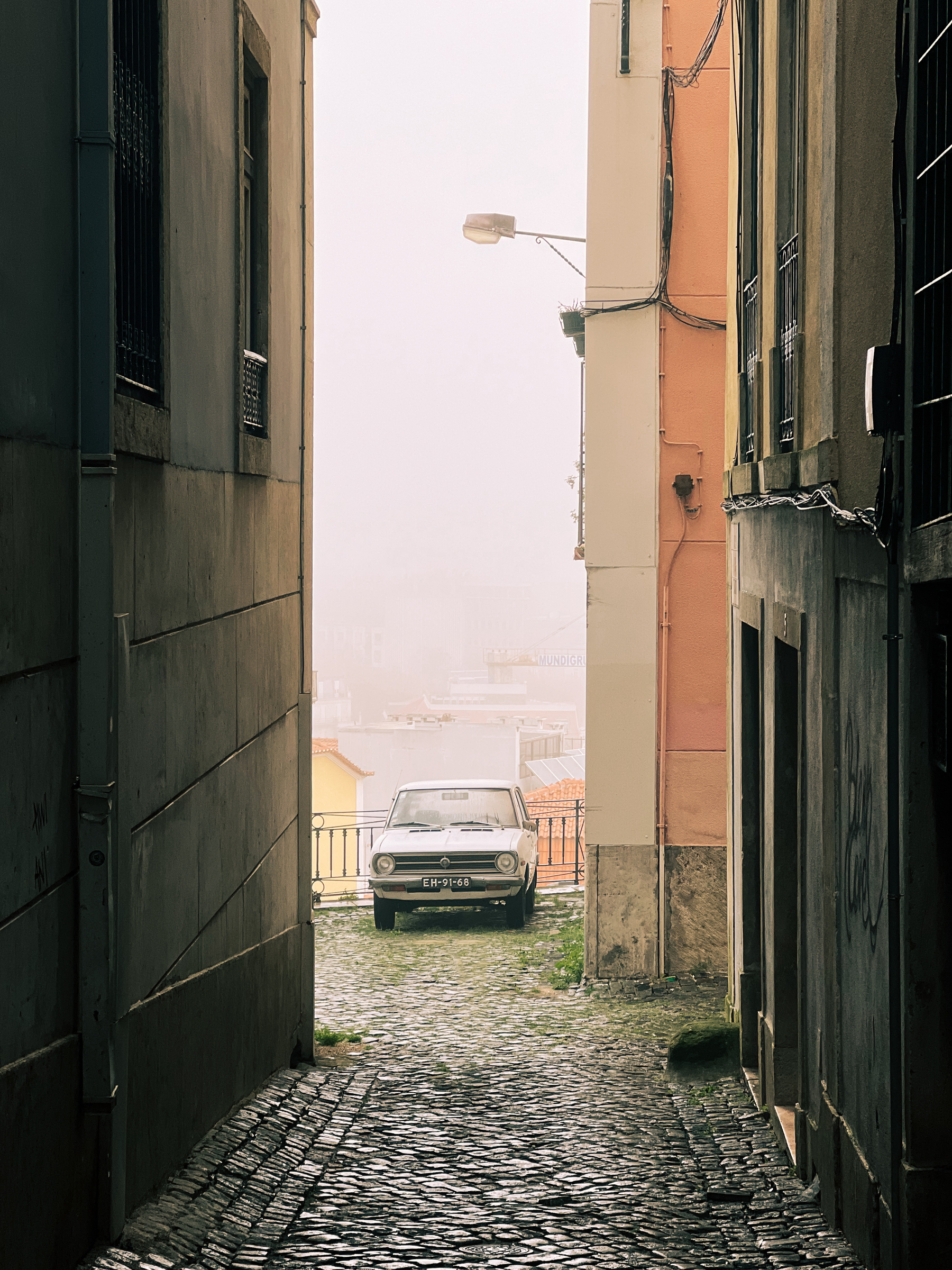 A narrow cobbled street bordered by buildings leads to a classic car parked at the end with buildings partly obscured by fog in the background.