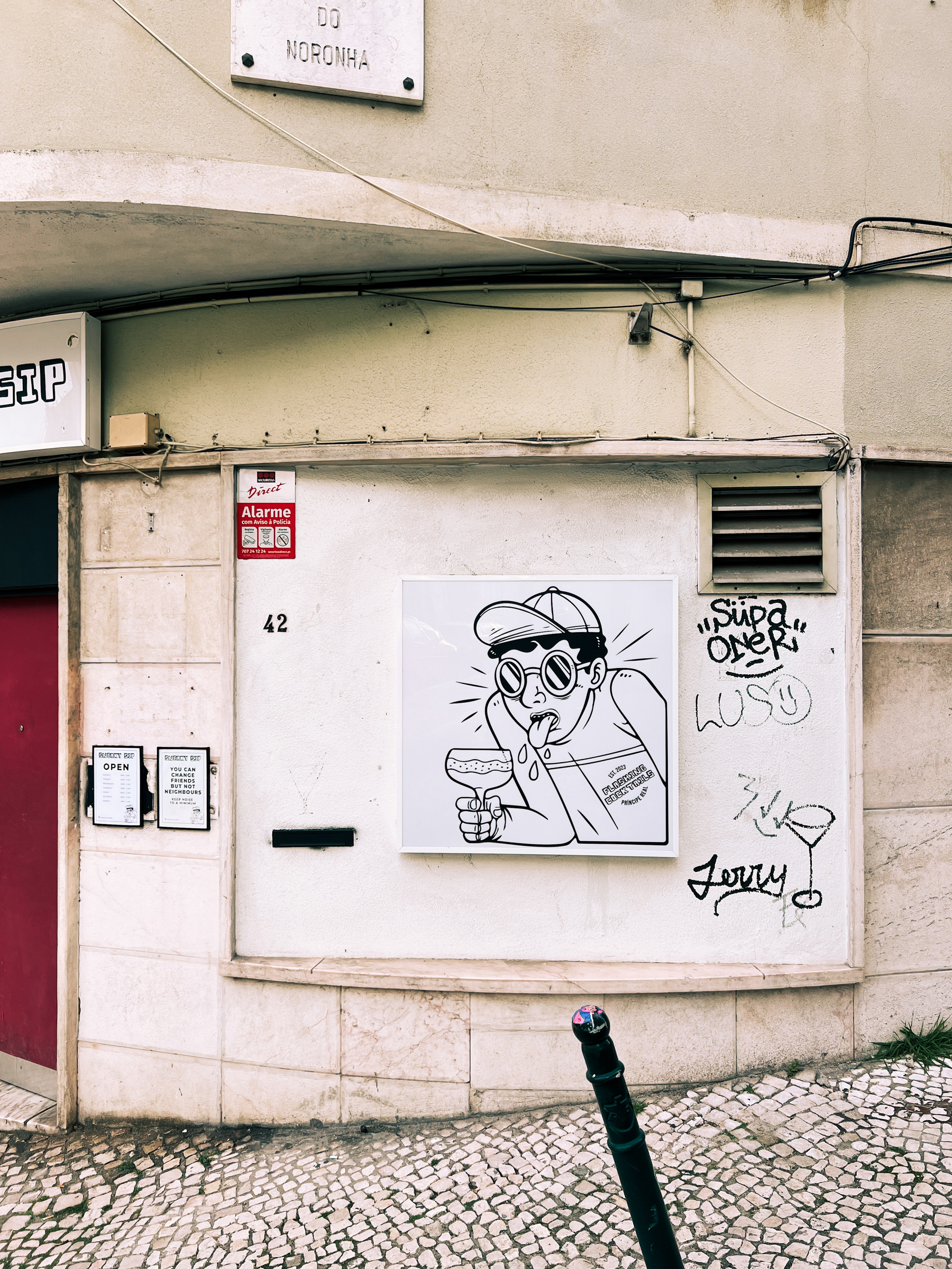 A street corner with a cartoon-style graffiti artwork on a wall, featuring a character wearing glasses and holding a cocktail. There are also signs, and some graffiti tags.