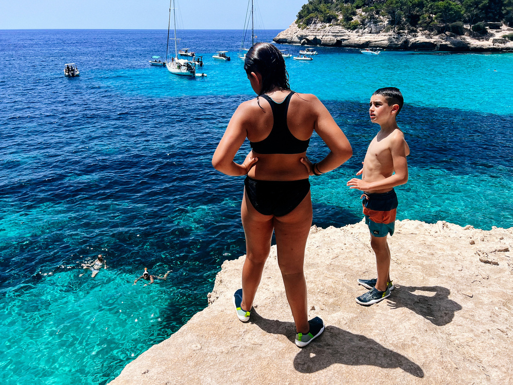 Two kids are standing on a rocky ledge overlooking clear, blue water with several boats floating in the distance. They are wearing swimwear and water shoes, and appear to be preparing for a jump into the sea. Swimmers are visible in the water.