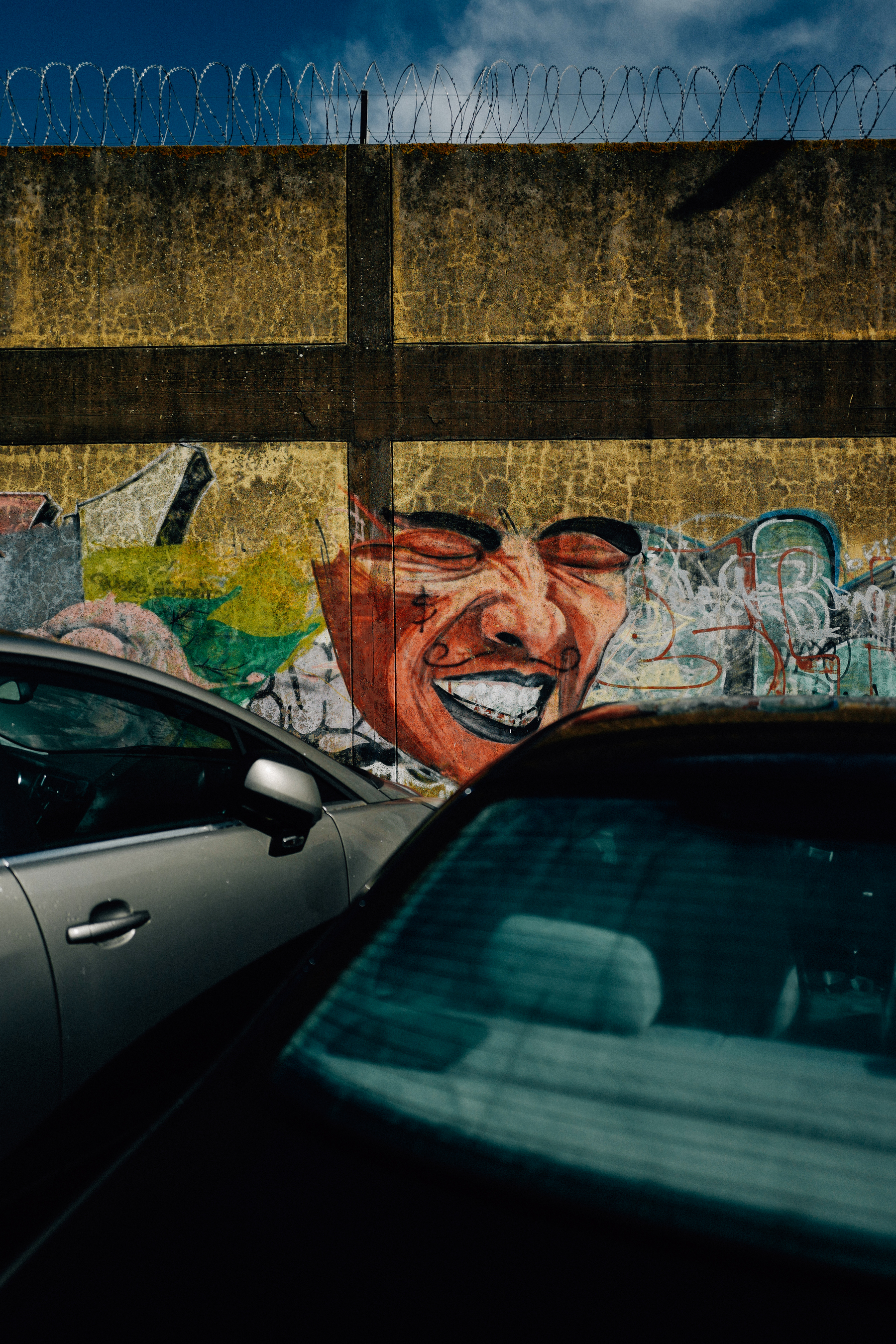 The image features a vibrant graffiti artwork of a face with an exaggerated expression on a wall, partially obscured by the top of two parked cars. The wall is topped with barbed wire, indicating a secured area. The overall tone is urban and gritty.