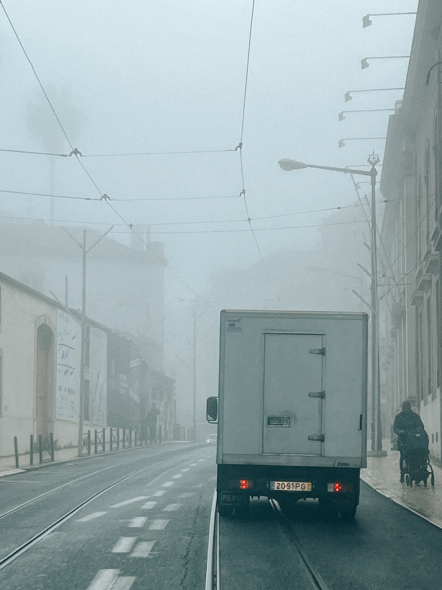 A foggy street scene with a delivery truck in the foreground and a person pushing a stroller in the background, with buildings and streetlamps lining the side of the road.