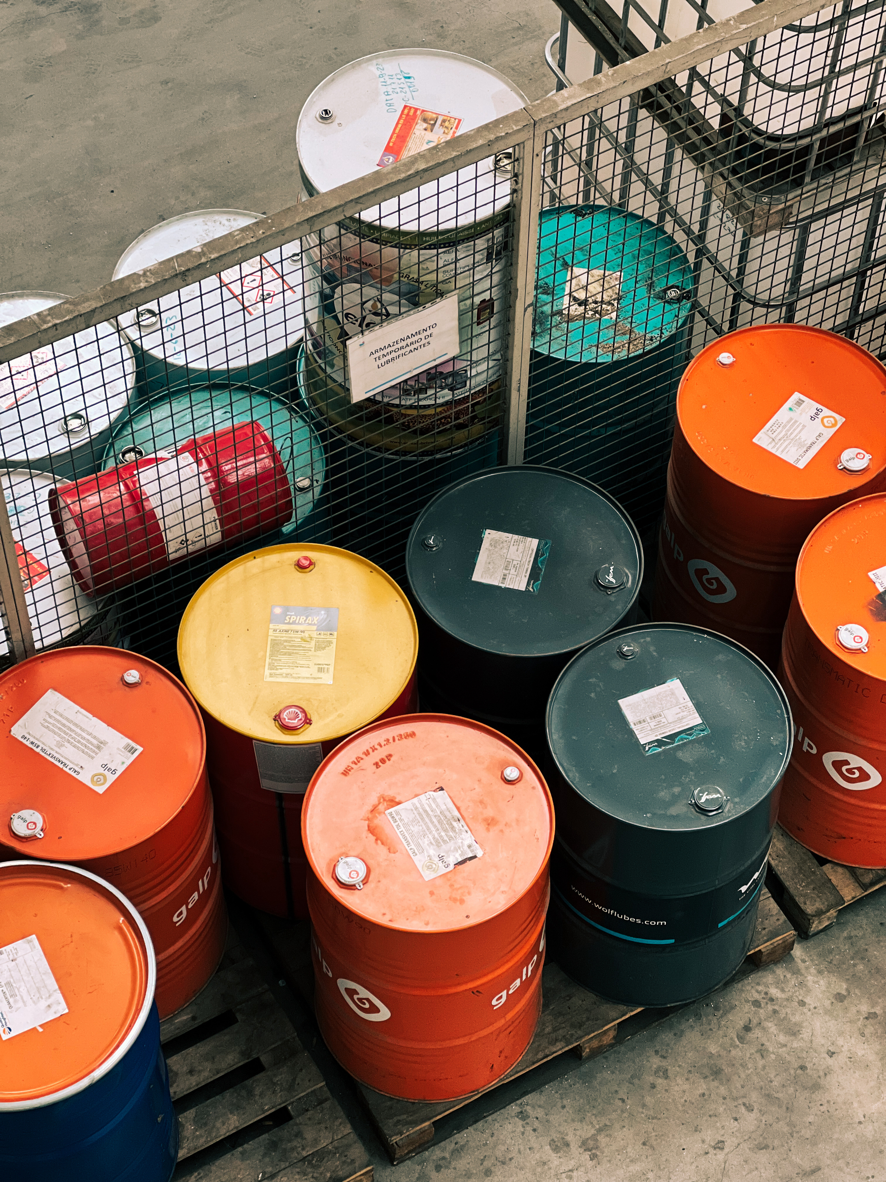 Industrial storage drums in various colors including red, yellow, blue, white, and black, stored within a metal cage on a concrete floor. Some barrels display labels and hazard signs.