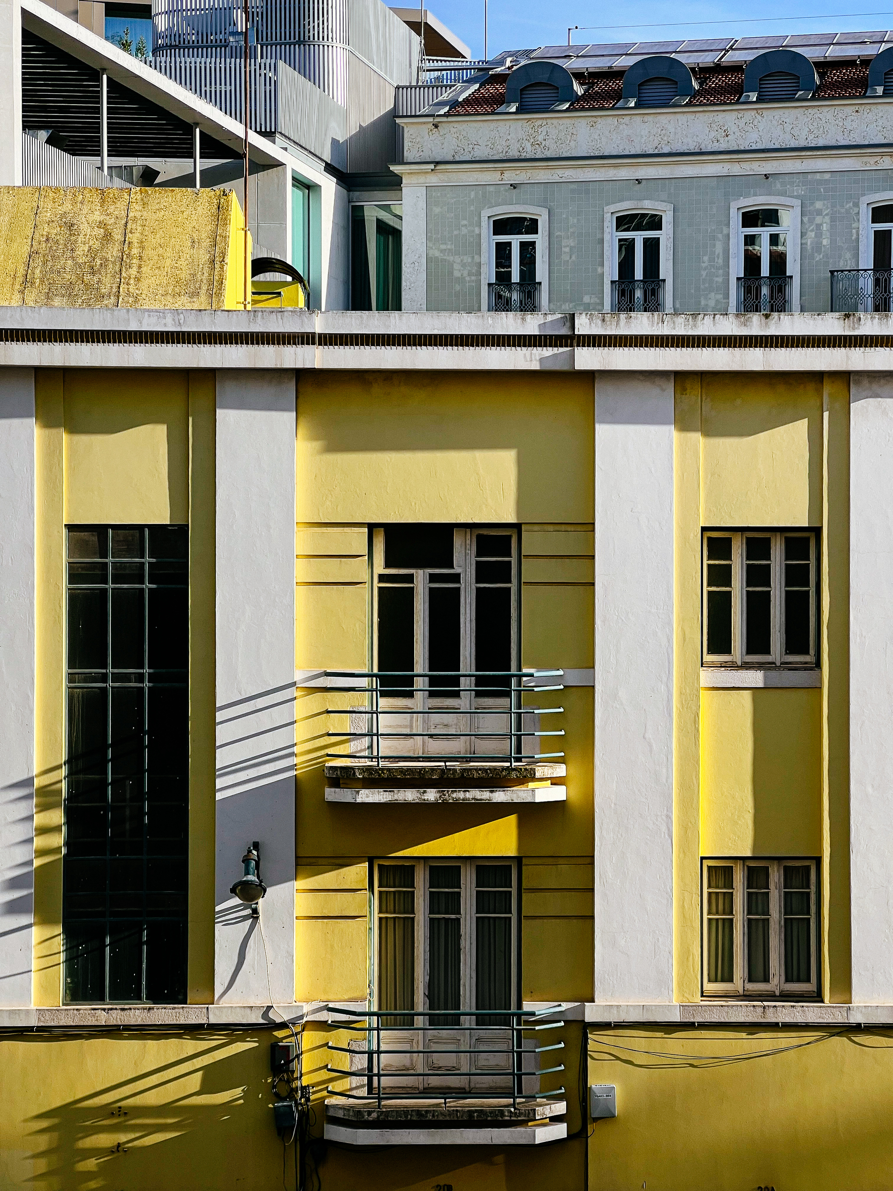A building facade with a contrasting architectural styles, featuring a yellow section with balconies and shadows cast on the walls, adjacent to a white building with classic windows and a rooftop terrace.