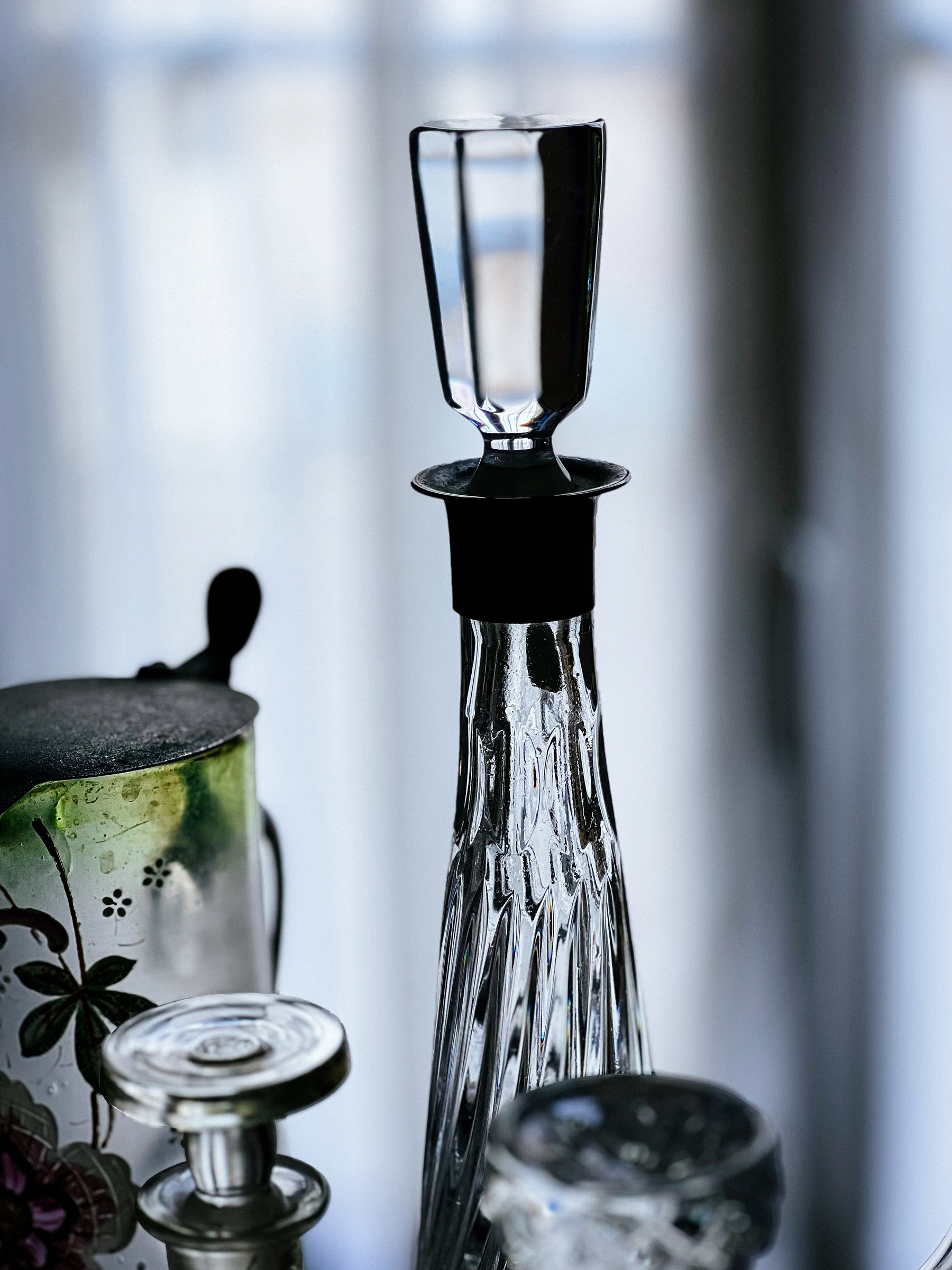 A close-up of a clear glass decanter with a stopper placed on top, surrounded by various glassware with intricate designs, set against a blurred background.