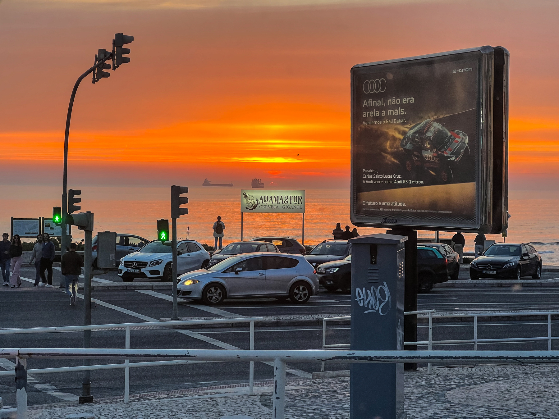 Sunset over a coastal road with pedestrians and cars, traffic lights, and a billboard featuring an Audi advertisement.